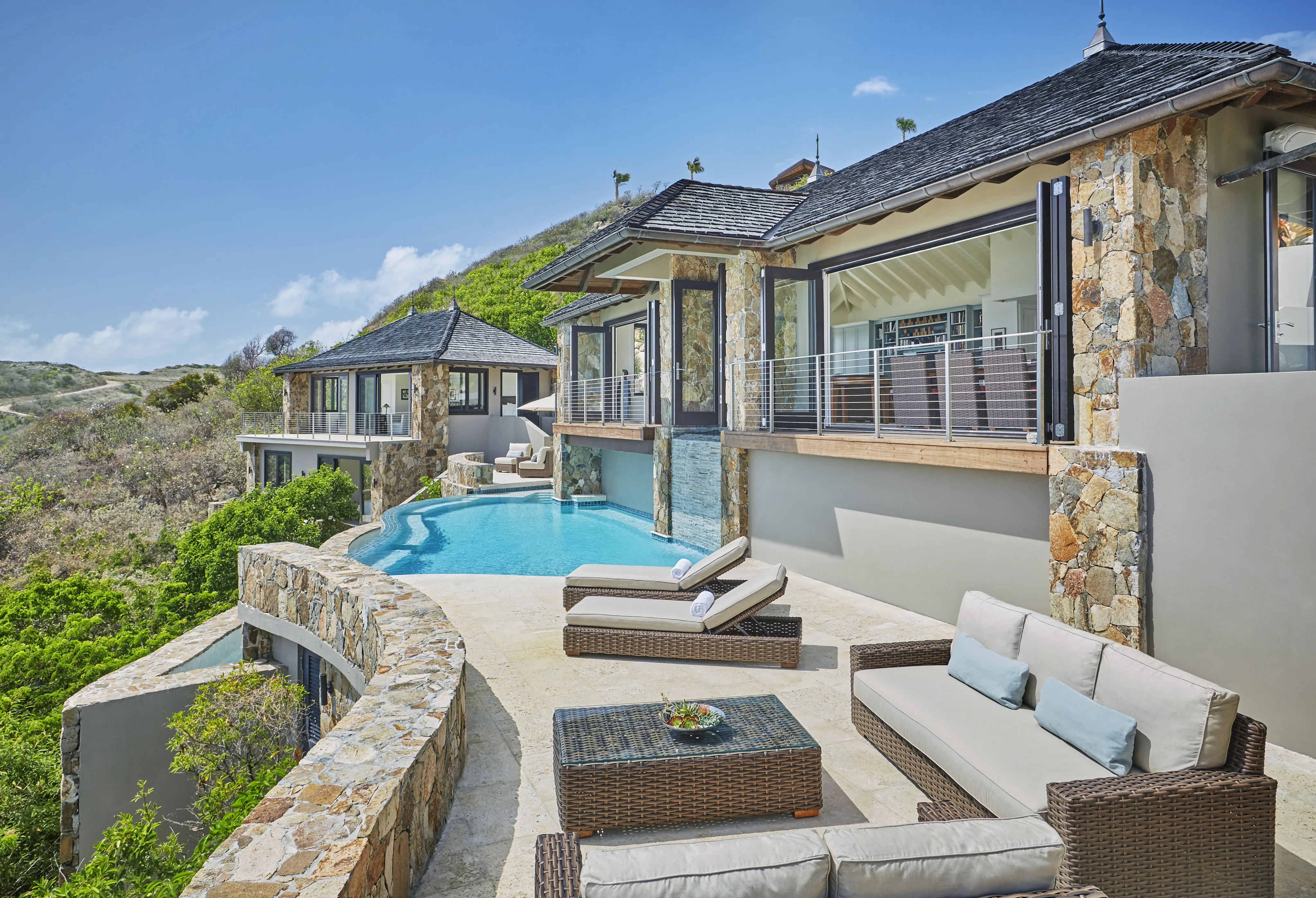 Cooke Lookout Villa is a luxurious hillside retreat with stone walls and an infinity pool that overlooks a lush green valley. The outdoor area includes sun loungers, a wicker coffee table, and a cushioned patio set under a clear blue sky. Adjoining rooms have large windows and sliding doors for breathtaking views.
