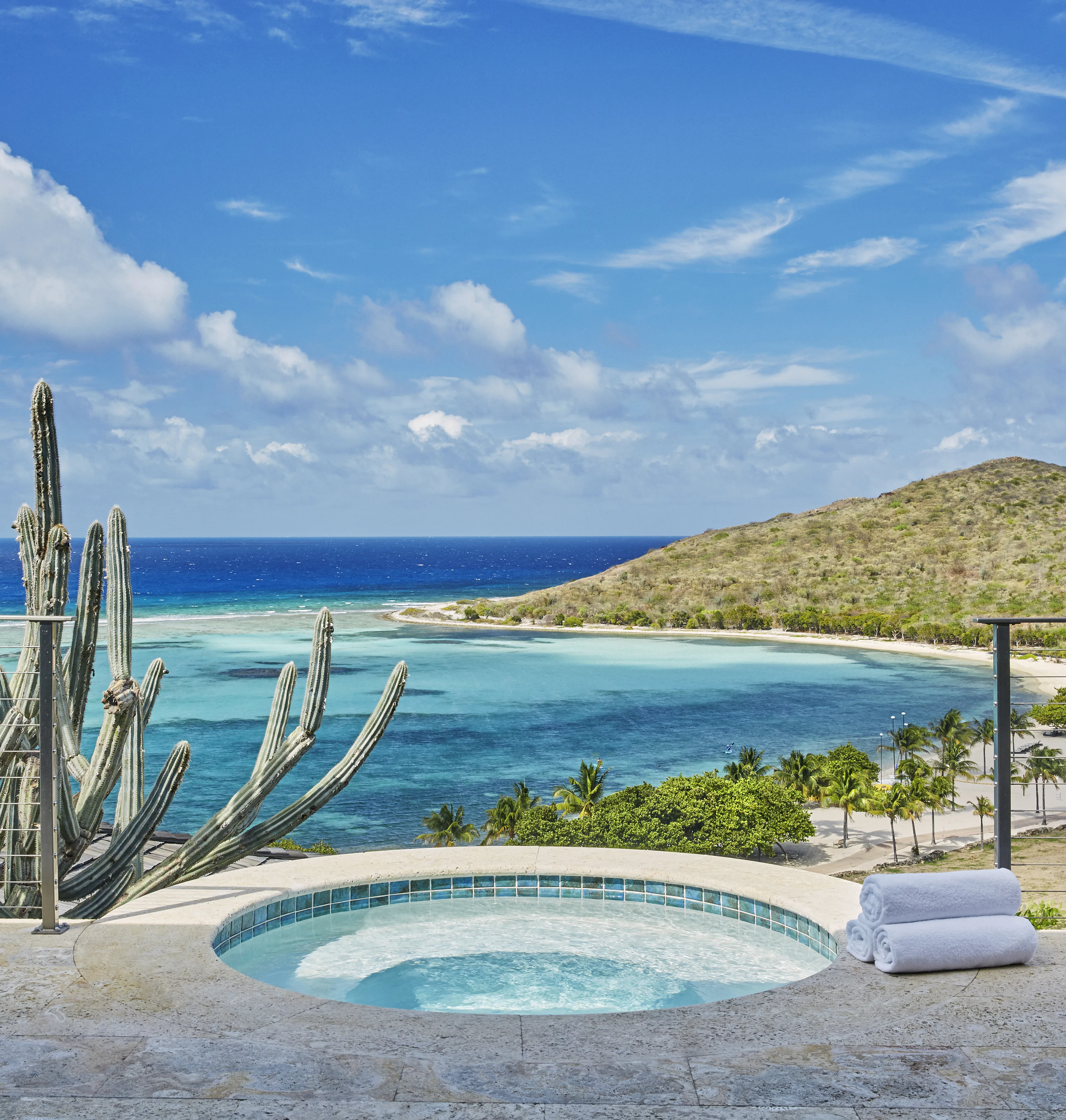 A serene view from the luxurious Cooke Lookout Villa pool overlooks a turquoise ocean bay with a small, hilly island in the background. Tall cacti are on the left, and the foreground features a clear blue hot tub with rolled white towels on the stone deck.