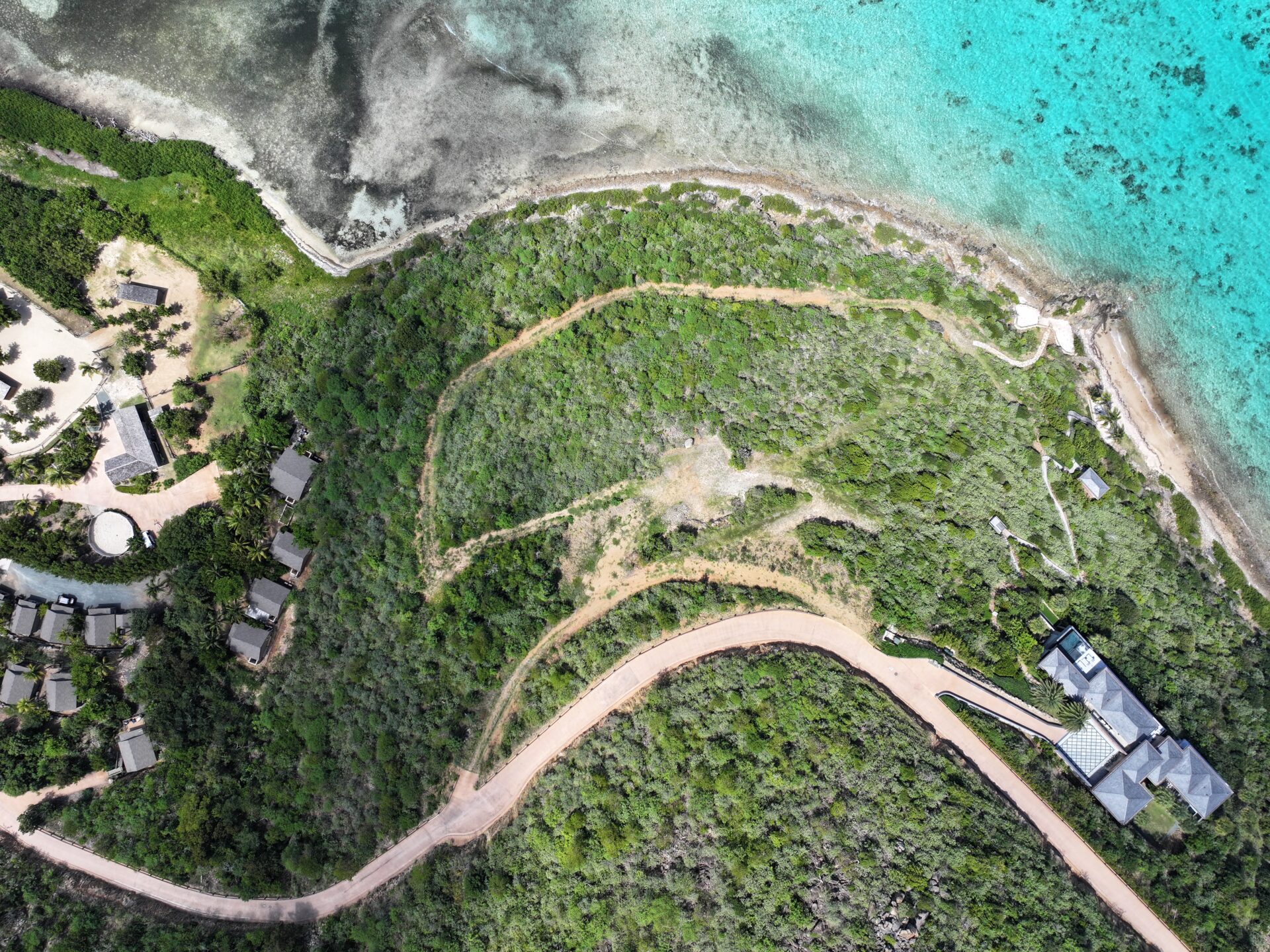 Aerial view of a coastal area with varying shades of water from turquoise to dark blue. The land is lush with green vegetation and winding roads leading to structures including houses and what appears to be an estate near the coastline.