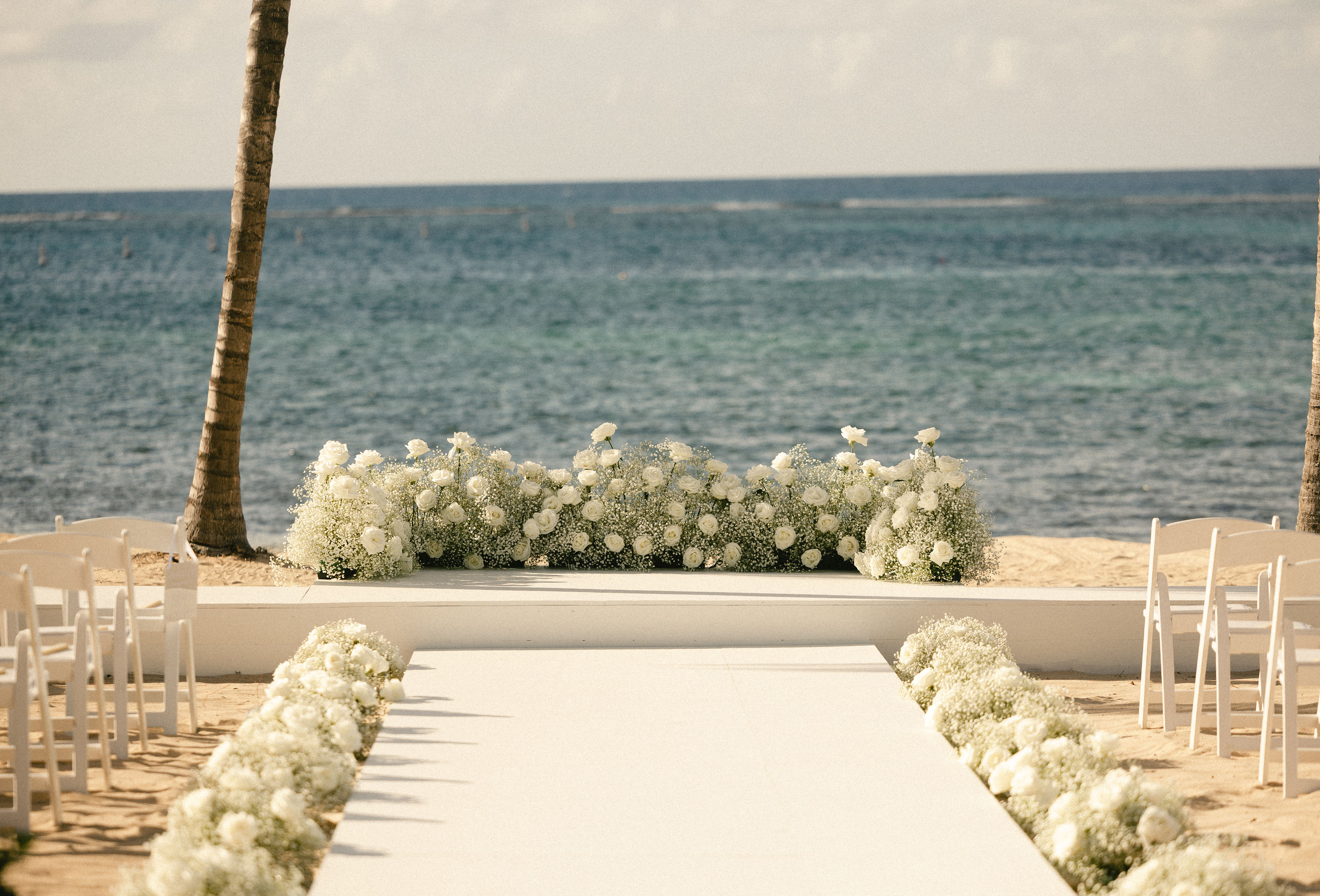 A beach wedding aisle lined with white flowers leads to an altar adorned in blooms, set against the ocean and a partly cloudy sky. White chairs flank the path, creating a scene of pure Wedding Bliss by the shore.