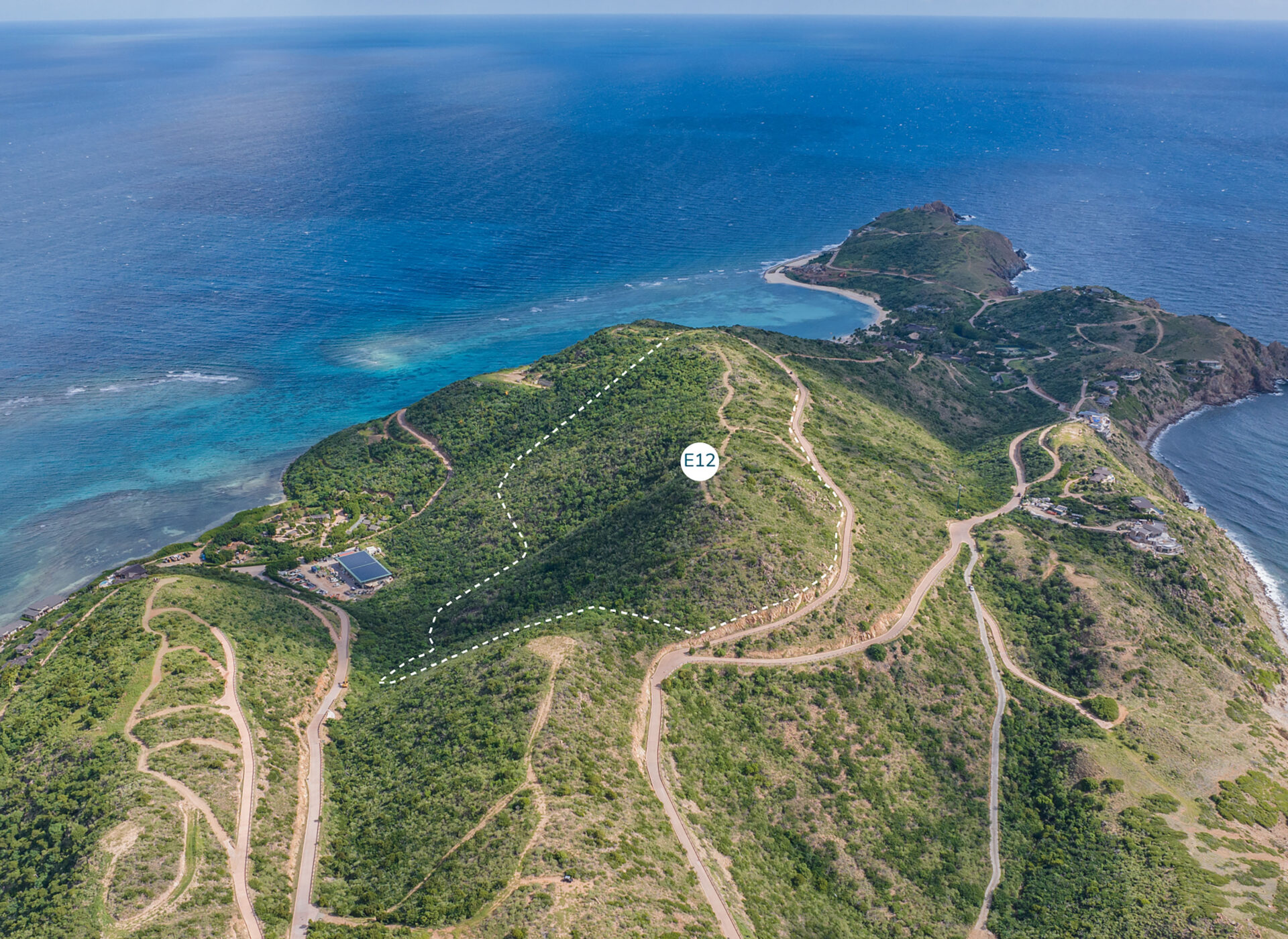 Aerial view of a hilly coastal landscape with winding roads and lush green vegetation. A marked area labeled "Estate Homesite E12" is visible on one of the hills. The serene ocean, with varying shades of blue, forms the backdrop. Sparse development is seen near the coast.