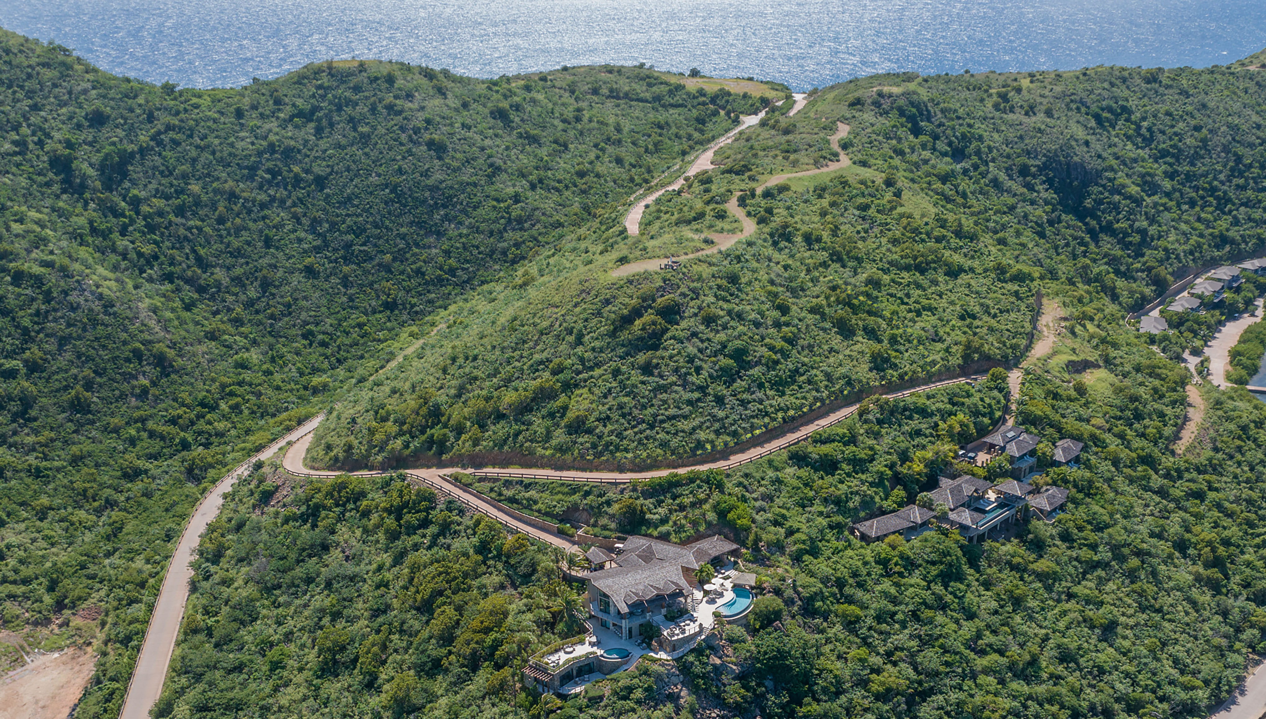 Aerial view of a lush, green, mountainous landscape with winding roads leading to a large, luxurious E5 estate homesite featuring multiple pools. The estate overlooks the ocean, which is visible in the background.