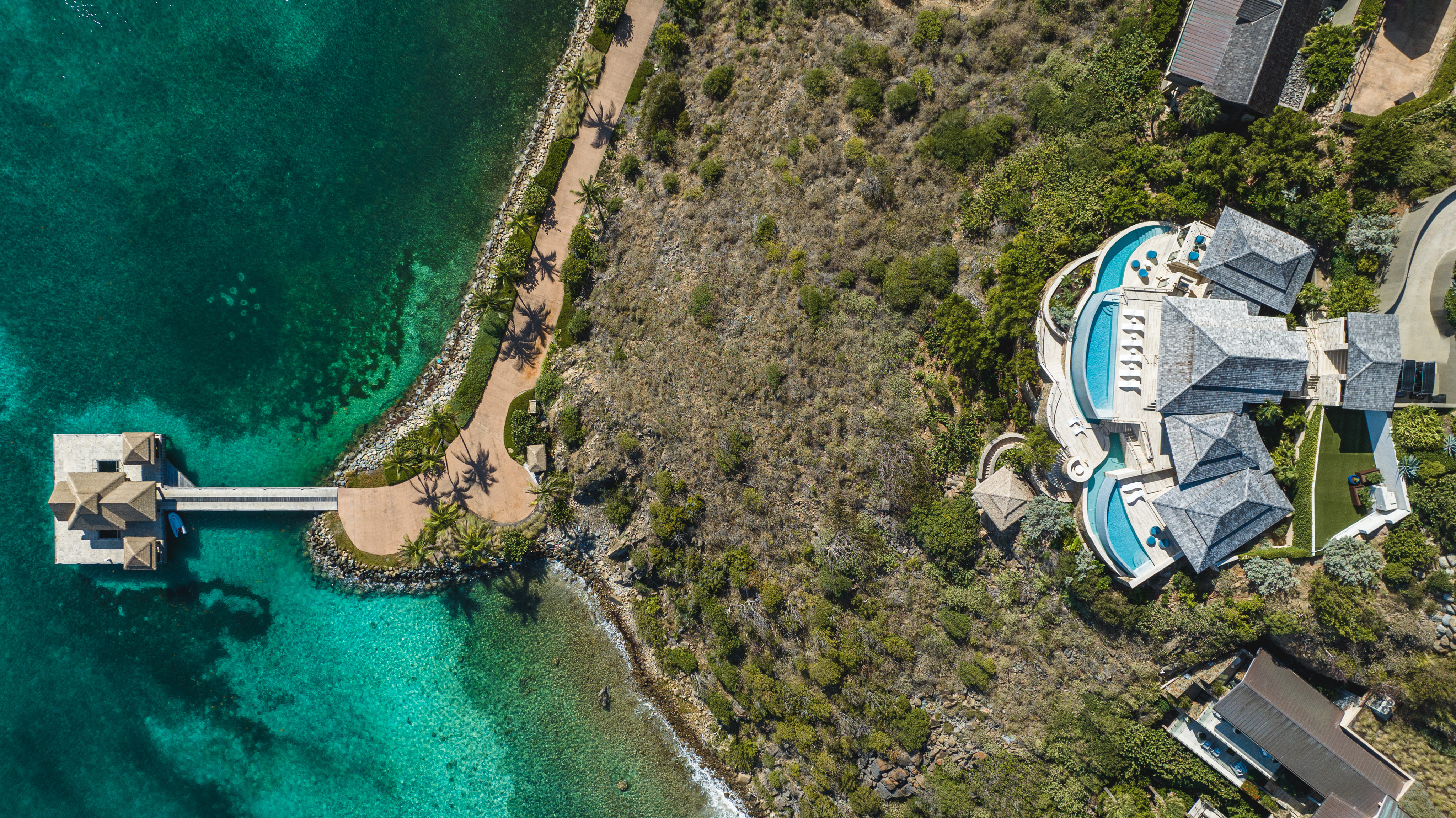 Aerial view of a luxury seaside Villa, aptly named Edge of Paradise, with a pool, surrounded by lush greenery and located next to a clear turquoise ocean. A pathway leads to a wooden pavilion over the water. The property and coastline are beautifully integrated with the natural landscape.