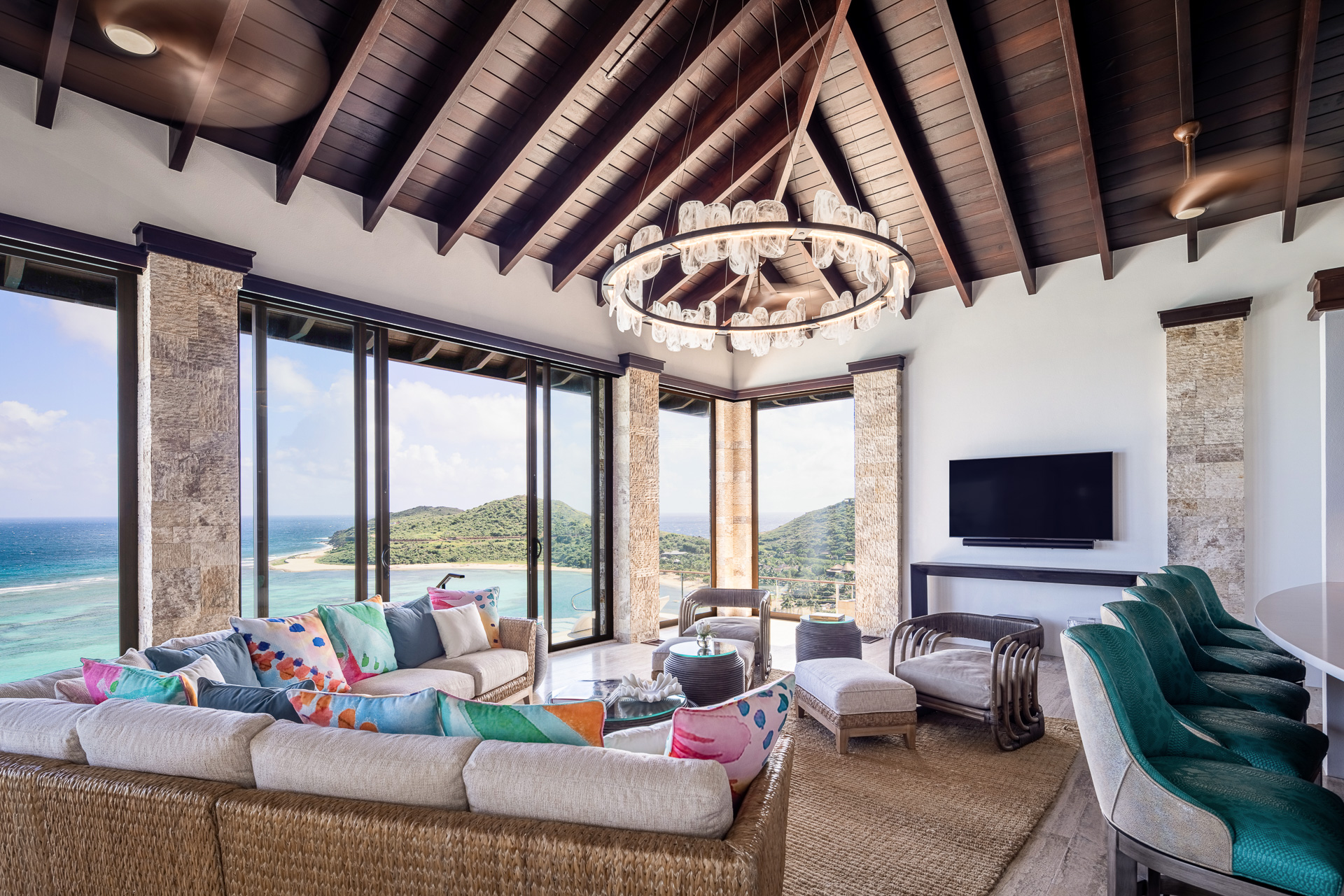 A luxurious, open-concept living room at Paradise Villa features a high, wooden beamed ceiling and large windows offering a panoramic view of the ocean and lush greenery outside. The room includes a wicker sofa set with colorful cushions, a large chandelier, and a mounted TV—the true edge of paradise.