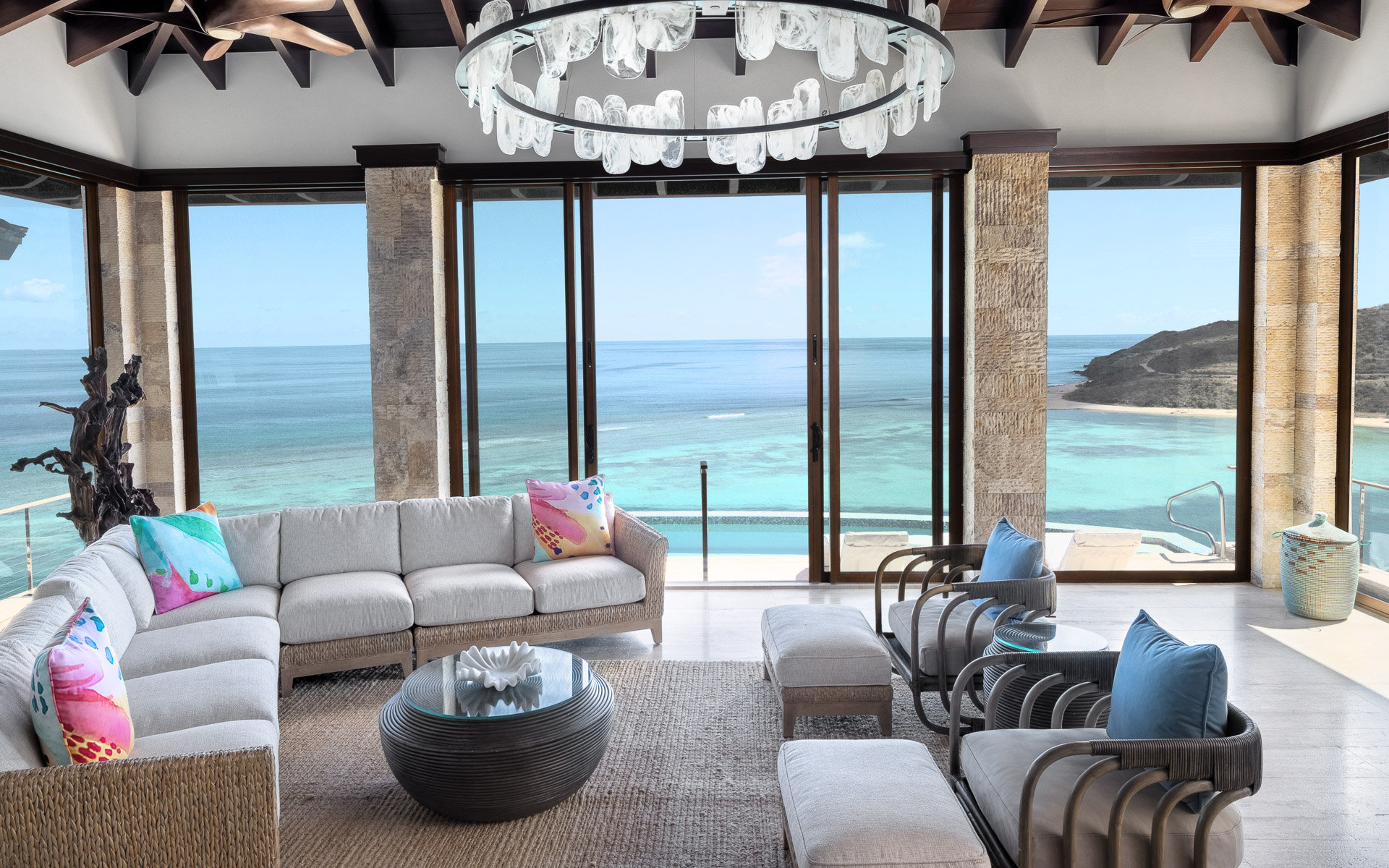 A modern living room in the Edge of Paradise Villa boasts large floor-to-ceiling windows offering a panoramic ocean view. It features a beige sectional sofa, wicker chairs with blue pillows, a glass coffee table, and a seashell chandelier. Light floods the room, highlighting the natural decor elements.
