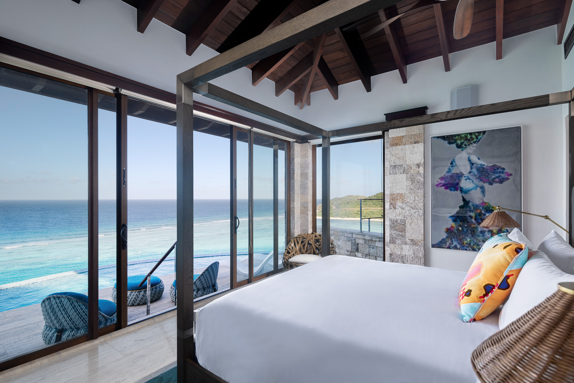 A luxurious bedroom at the Edge of Paradise features a large bed adorned with colorful pillows and a canopy frame. The room boasts floor-to-ceiling windows offering a stunning ocean view, while outside, a seating area on the deck overlooks the blue water and distant shoreline under a high wooden ceiling.