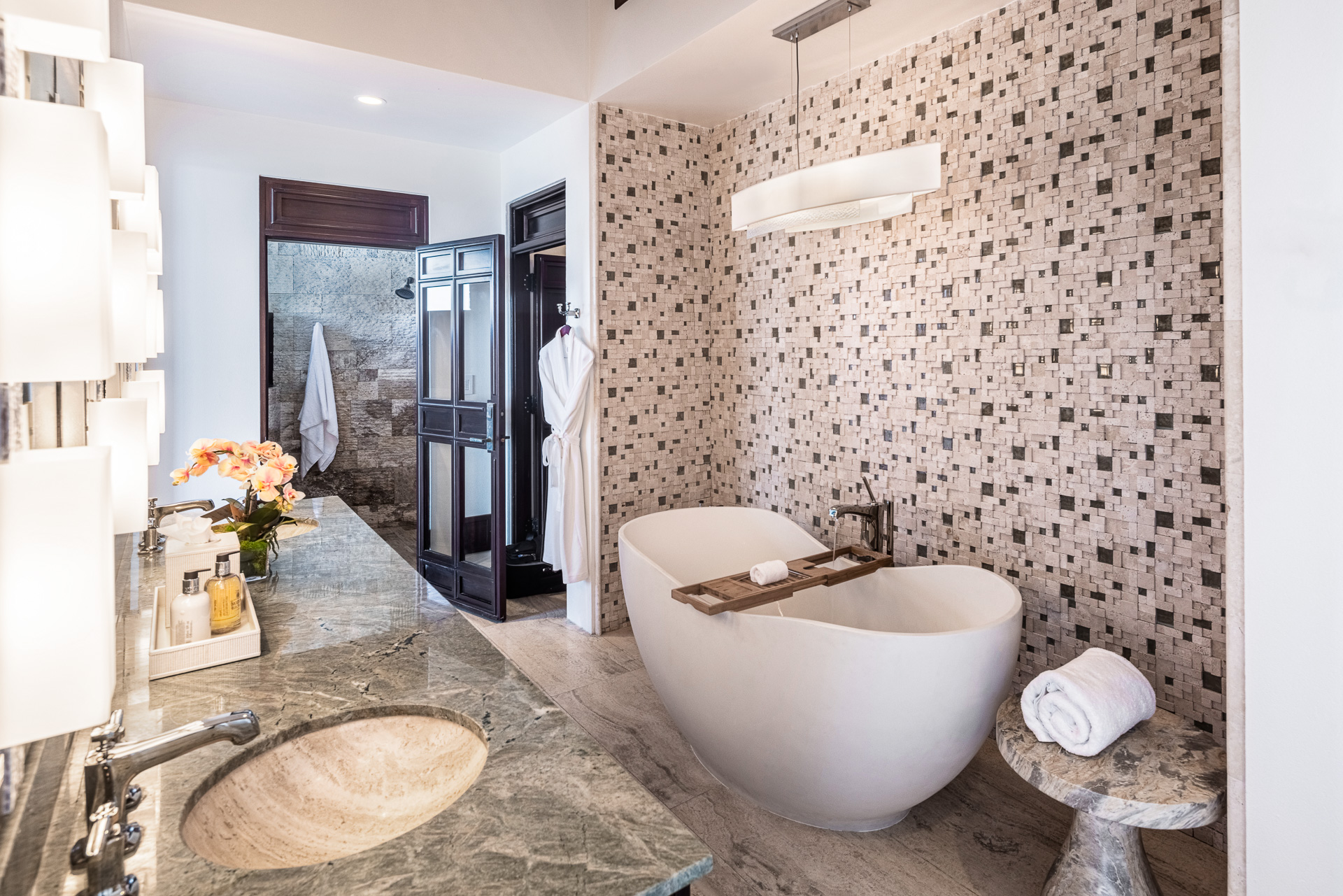 A modern bathroom in Paradise Villa features a large white freestanding bathtub, stone-tiled wall, marble countertop with a sink, vanity mirror, and glass-enclosed shower. A wooden tray rests on the tub, while two white robes hang near the shower—a true Edge of Paradise.