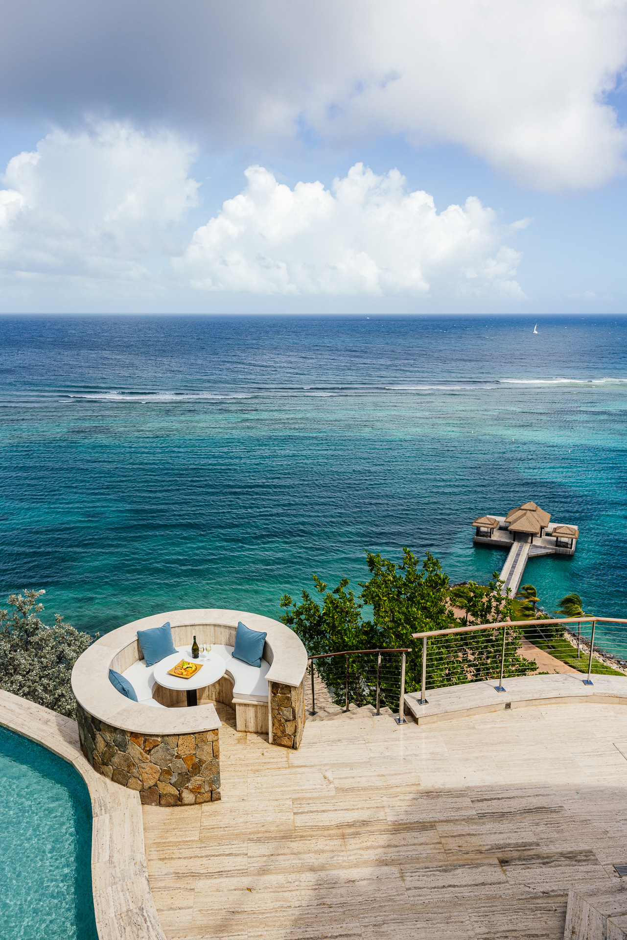 A tranquil seaside view at the Edge of Paradise villa features a luxurious poolside seating area with turquoise cushions, overlooking a vast expanse of blue ocean. A wooden pier with a thatched hut extends into the water. Lush greenery and clear skies enhance the serene ambiance.