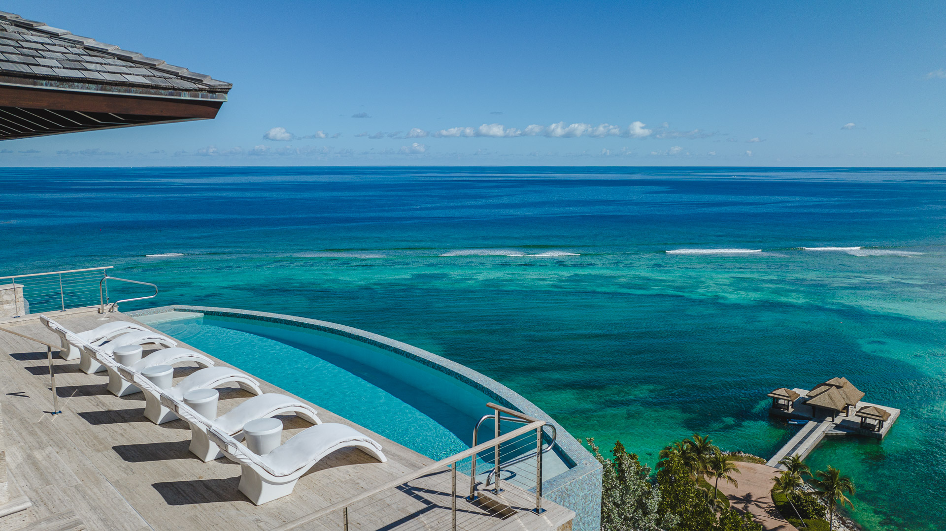 A luxurious infinity pool with white loungers at the Villa overlooks the vast, serene ocean, embodying the Edge of Paradise. The water transitions from light to deep blue, and a small wooden pier extends into the clear water. The scene is tranquil, with a bright, clear sky overhead.