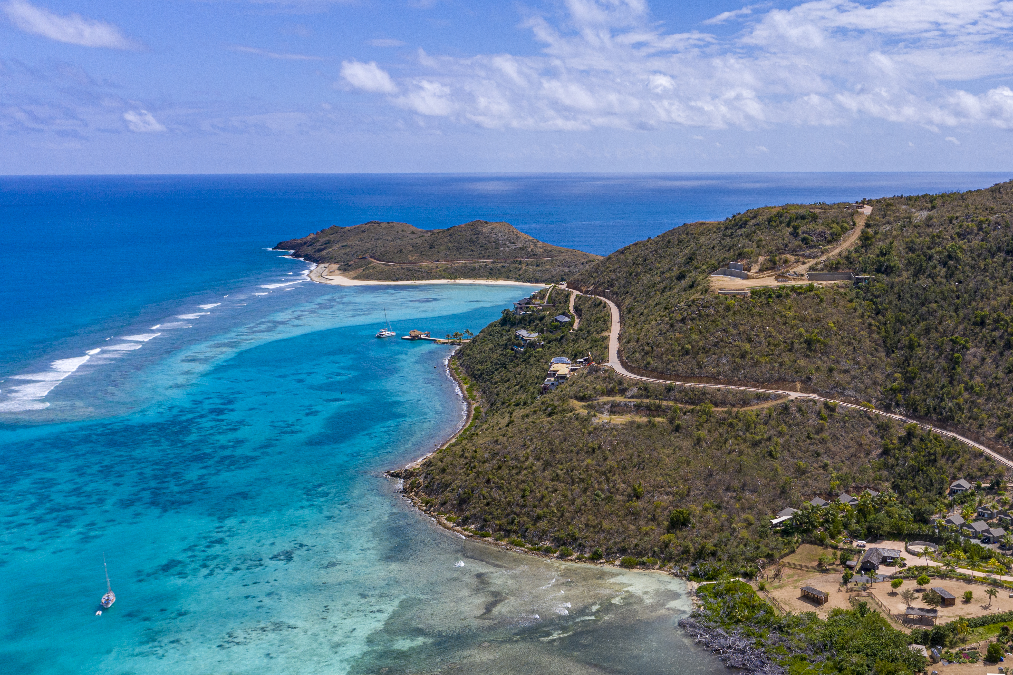 Aerial view of a winding coastal road on a hilly island, surrounded by turquoise waters and coral reefs. The coastline features several small buildings, lush greenery, and an estate nestled near the shore. A lone sailboat is anchored nearby under a partly cloudy sky.