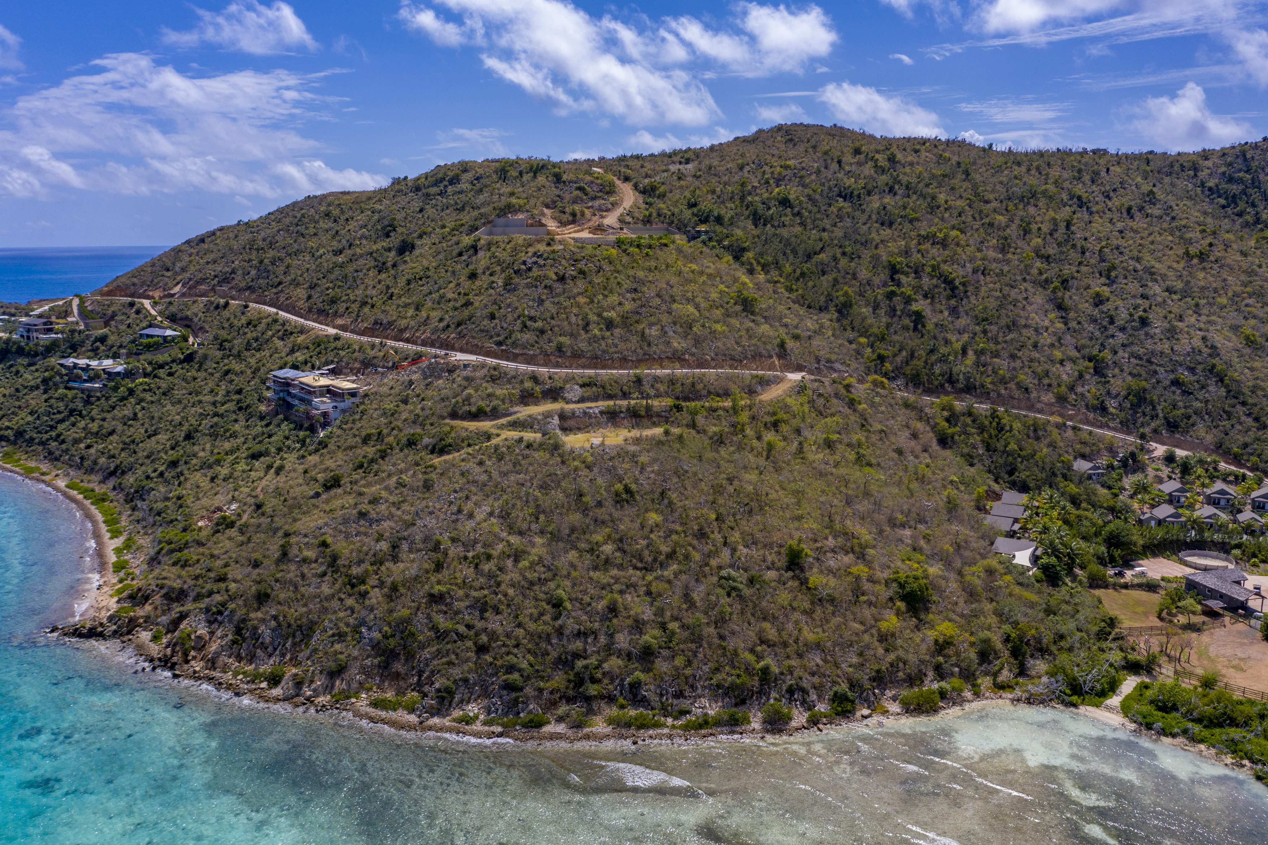 An aerial view of a coastal hillside covered in green and brown vegetation reveals winding roads and a few scattered homesites. The clear, turquoise waters of the ocean border the shoreline on the left, while the partly cloudy sky stretches overhead.