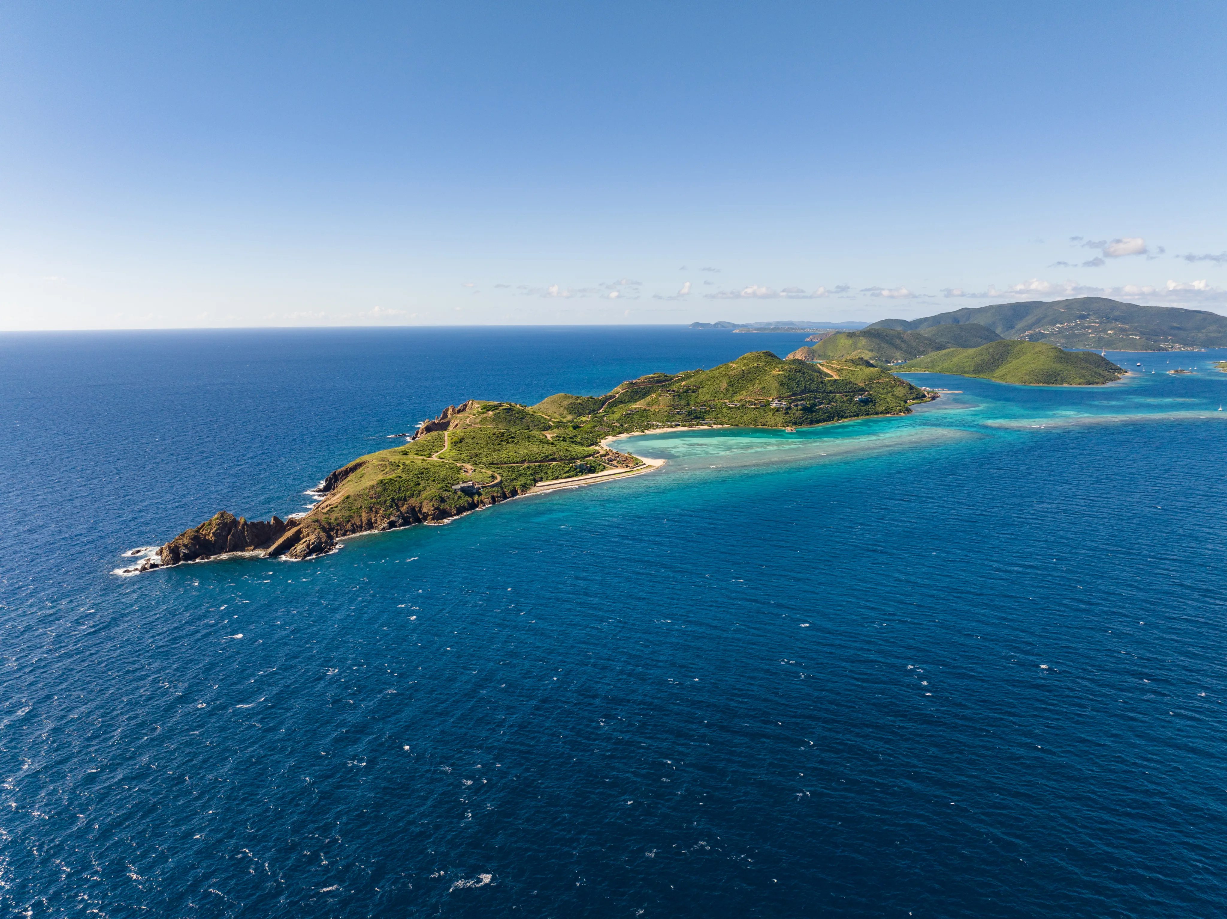 Aerial view of a picturesque island surrounded by turquoise waters and lush green hills, resembling the ideal British Virgin Islands resort. White sandy beaches line the coast, and the horizon stretches across a clear, blue sky. Small islets are visible in the distance.