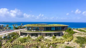 Halo Villa, a modern, two-story house with a curved design and a green roof, is set against the backdrop of the ocean. The house features large windows and a lush, landscaped garden. Palm trees and clear blue skies complete the coastal view for this stunning abode.