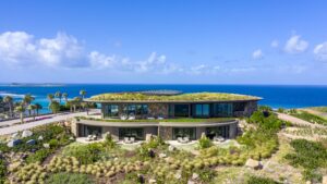 Halo Villa, a modern, two-story house with a curved design and a green roof, is set against the backdrop of the ocean. The house features large windows and a lush, landscaped garden. Palm trees and clear blue skies complete the coastal view for this stunning abode.