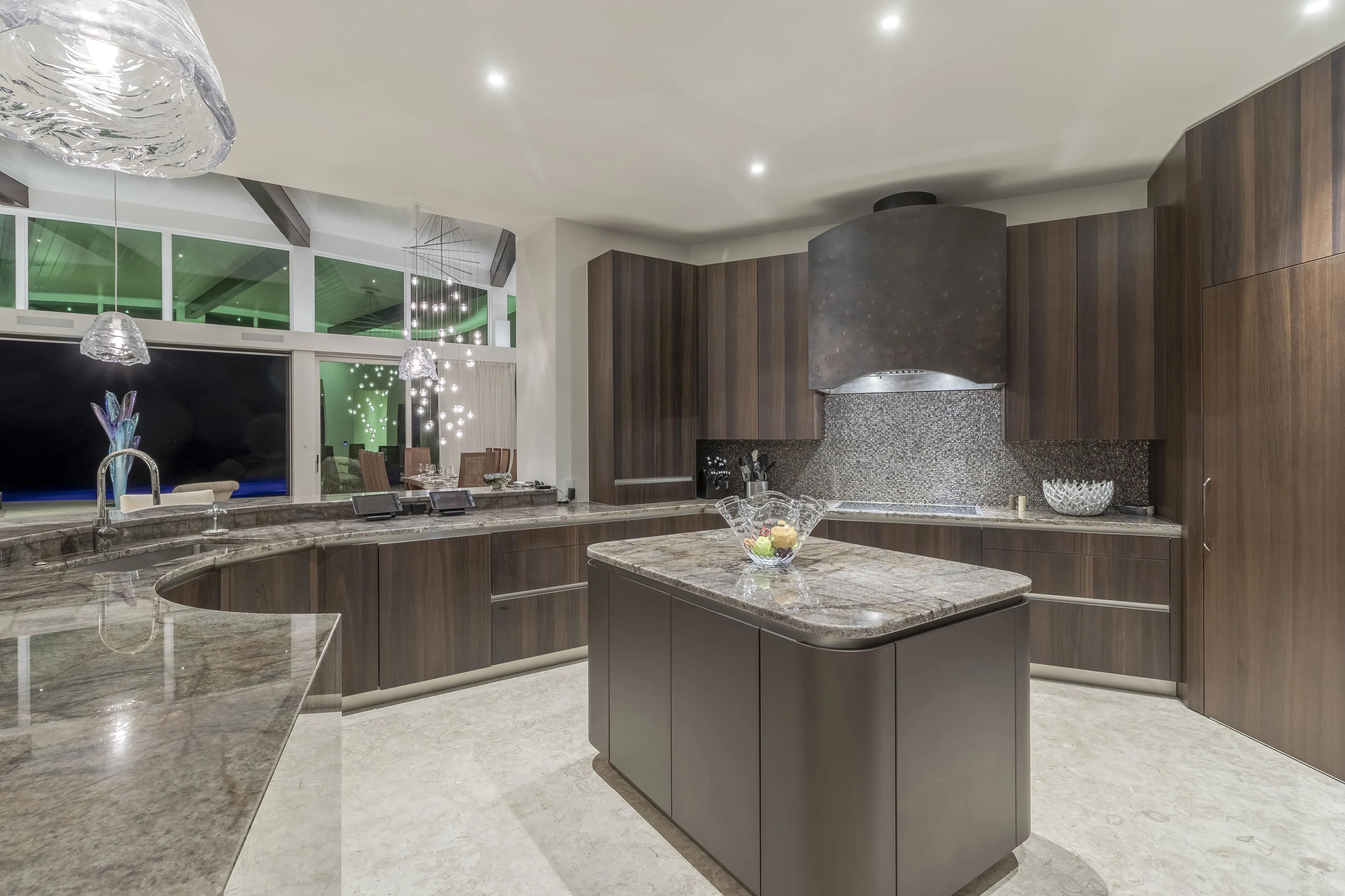 Modern kitchen with sleek dark wood cabinetry and a large marble central island, fitting perfectly within the Halo Villa. The countertops are also marble, and there is a distinctive large black range hood above the stove. A bowl of fruit decorates the island. Ambient lighting creates a cozy atmosphere.