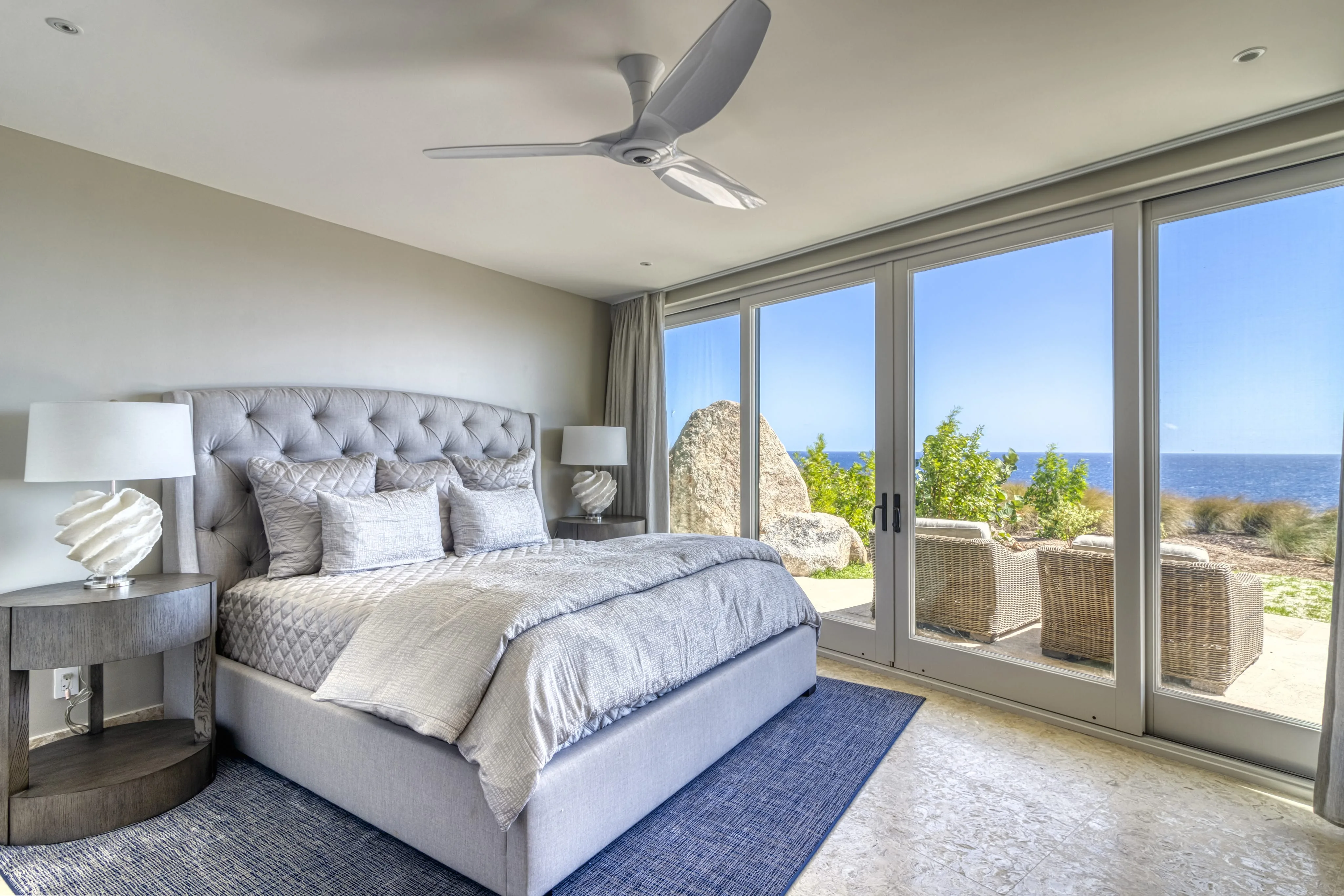 A modern bedroom in Halo Villa features a large bed with a tufted headboard, flanked by two bedside tables with lamps. The room has a ceiling fan and opens up to a patio with wicker furniture, overlooking a scenic ocean view through large glass doors.