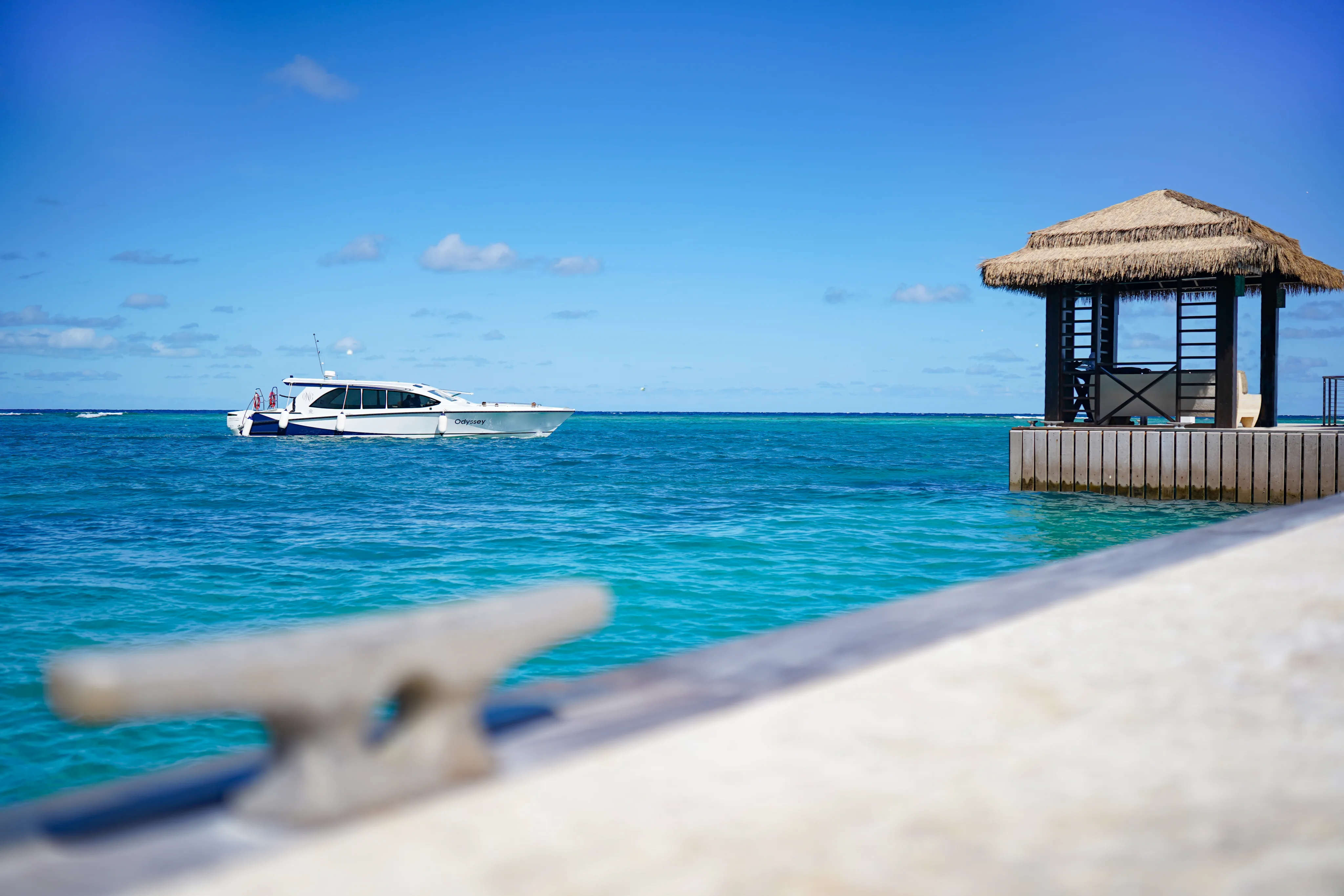 A boat floats on clear blue water under a sunny sky, with a small hut with a thatched roof on the right side of the image. A blurred foreground object, likely part of a dock or waterfront structure, adds to the tranquil scene, inviting you to stay and enjoy this idyllic paradise.