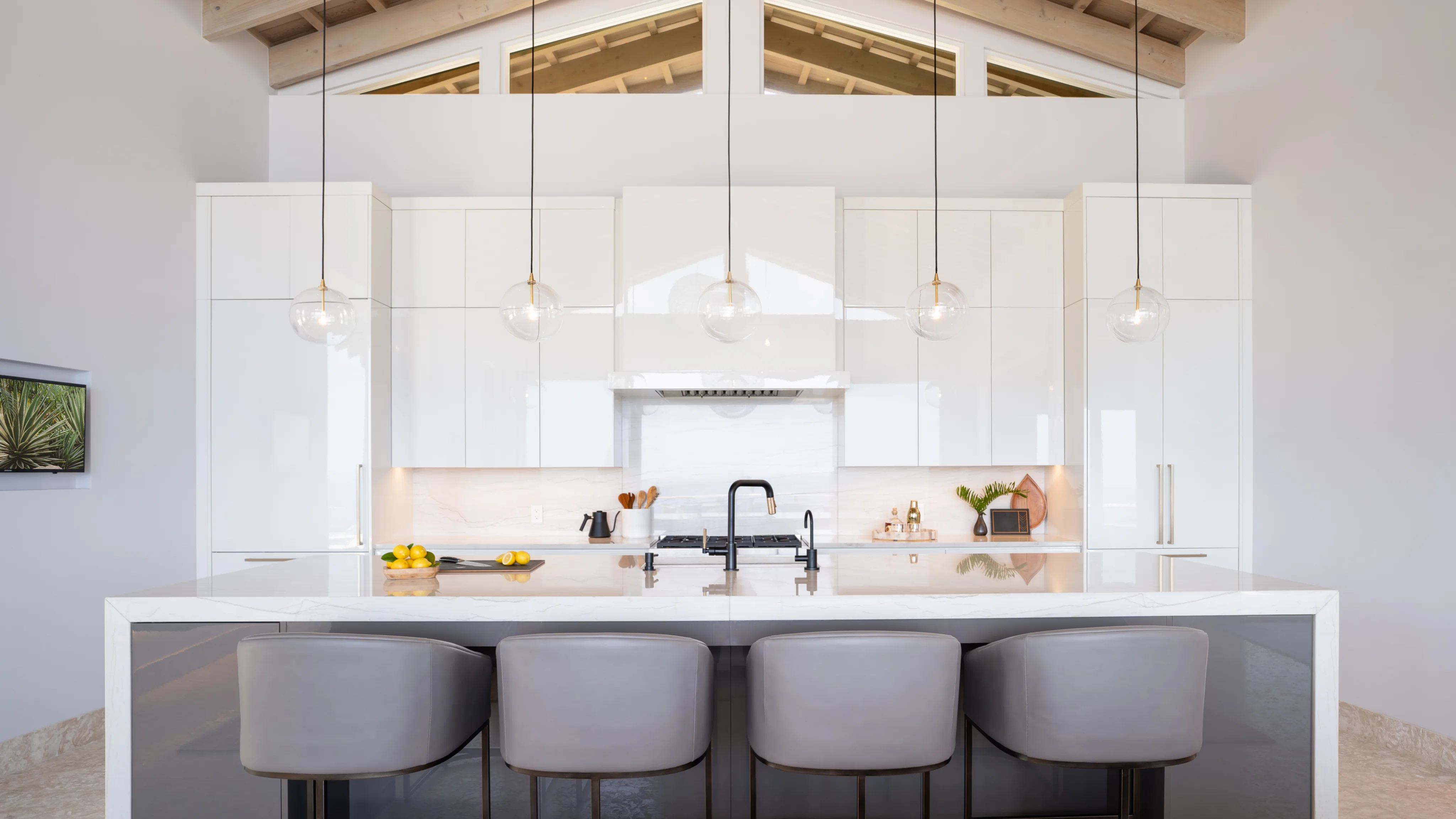 A modern villa kitchen features a sleek white design with high ceilings and open beams. A large island with a marble countertop and four gray barstools occupies the center. Pendant lights hang above the island, resembling graceful hummingbirds in flight. The backdrop includes cabinets, a stove, and a range hood.