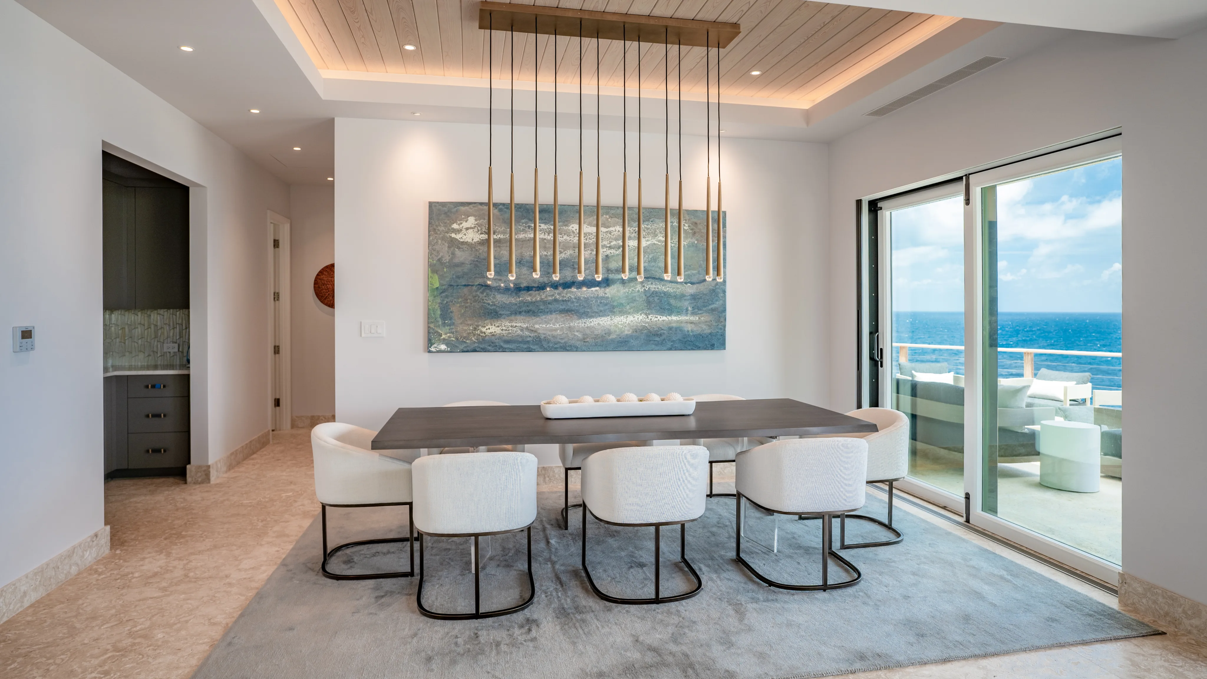 A modern dining room in the villa features a long table with eight white chairs arranged around it. Above the table hangs a linear chandelier with multiple pendant lights. A large, abstract painting decorates the wall, and sliding glass doors open to a view of the ocean where hummingbirds often visit.