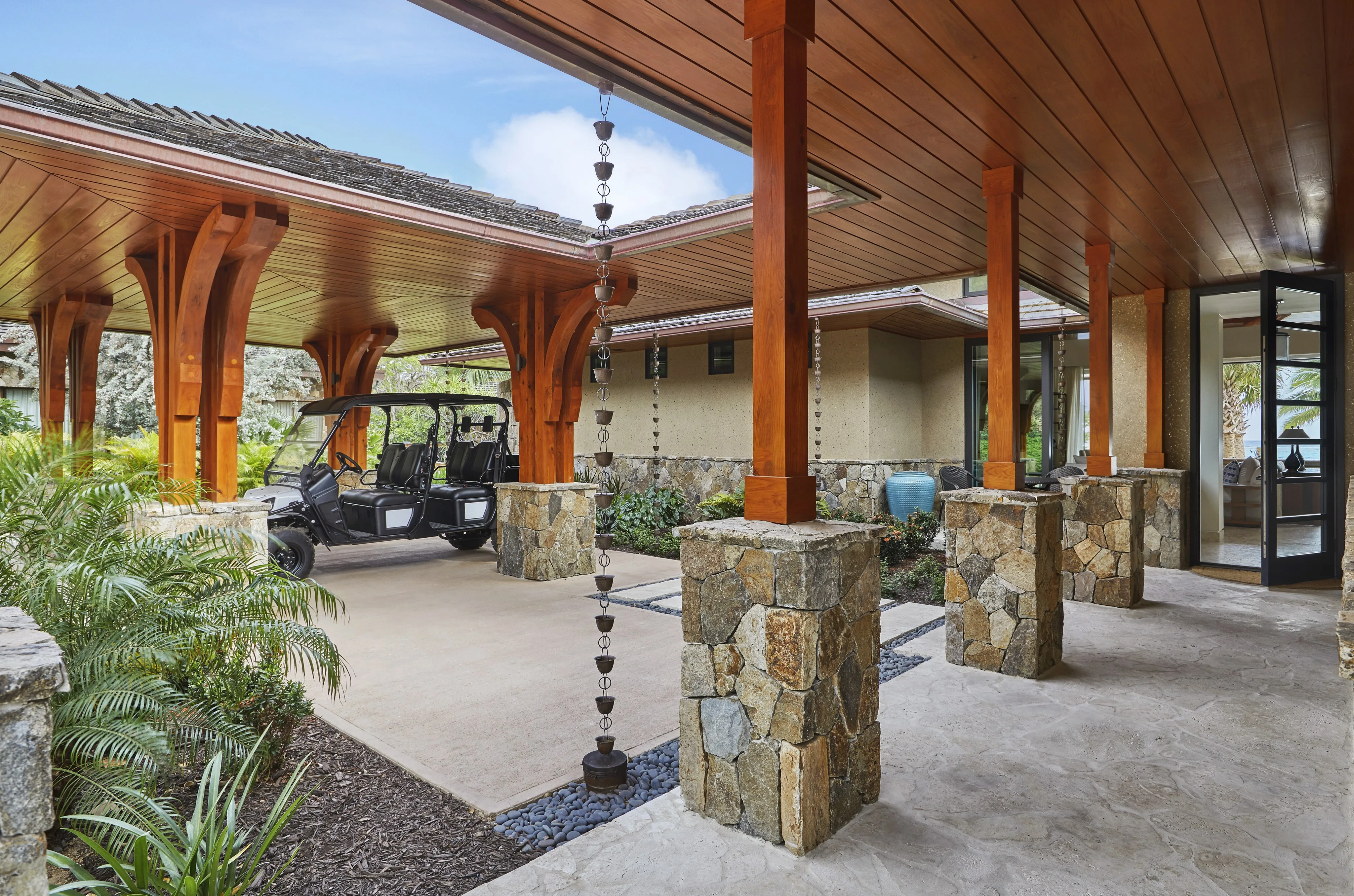 Cheemaun Villa boasts a luxurious open carport with wooden beams and stone pillars. A black golf cart rests under the elegant roof. There are hanging rain chains, tropical plants, and a clear view inside the modern house through large windows. The sky is blue with scattered clouds.