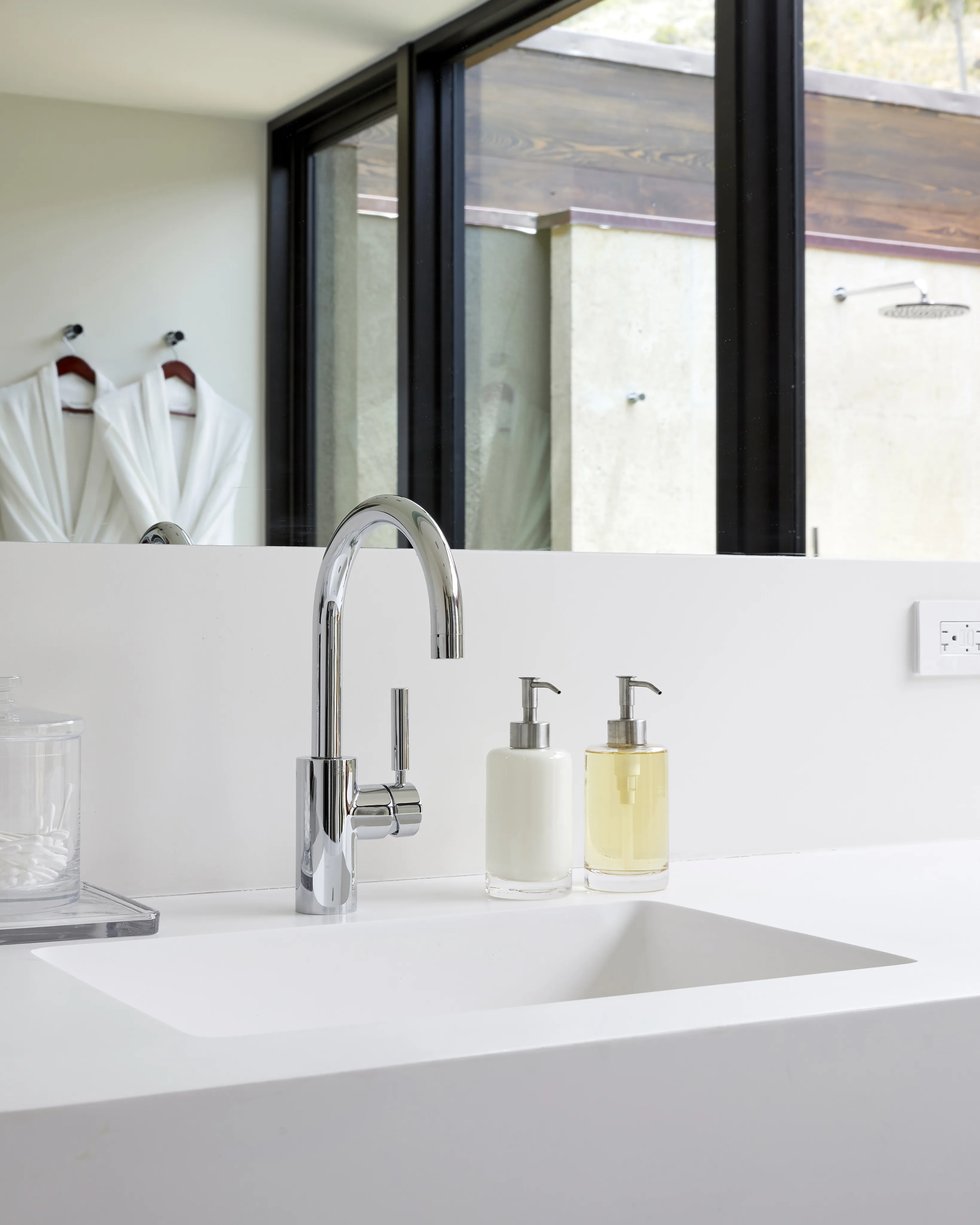 A modern bathroom in a beach house features a sleek, white countertop with a built-in sink. A chrome faucet is mounted on the countertop alongside two soap dispensers, one with white liquid and the other with yellow. In the background, a glass wall reveals two white robes hanging.