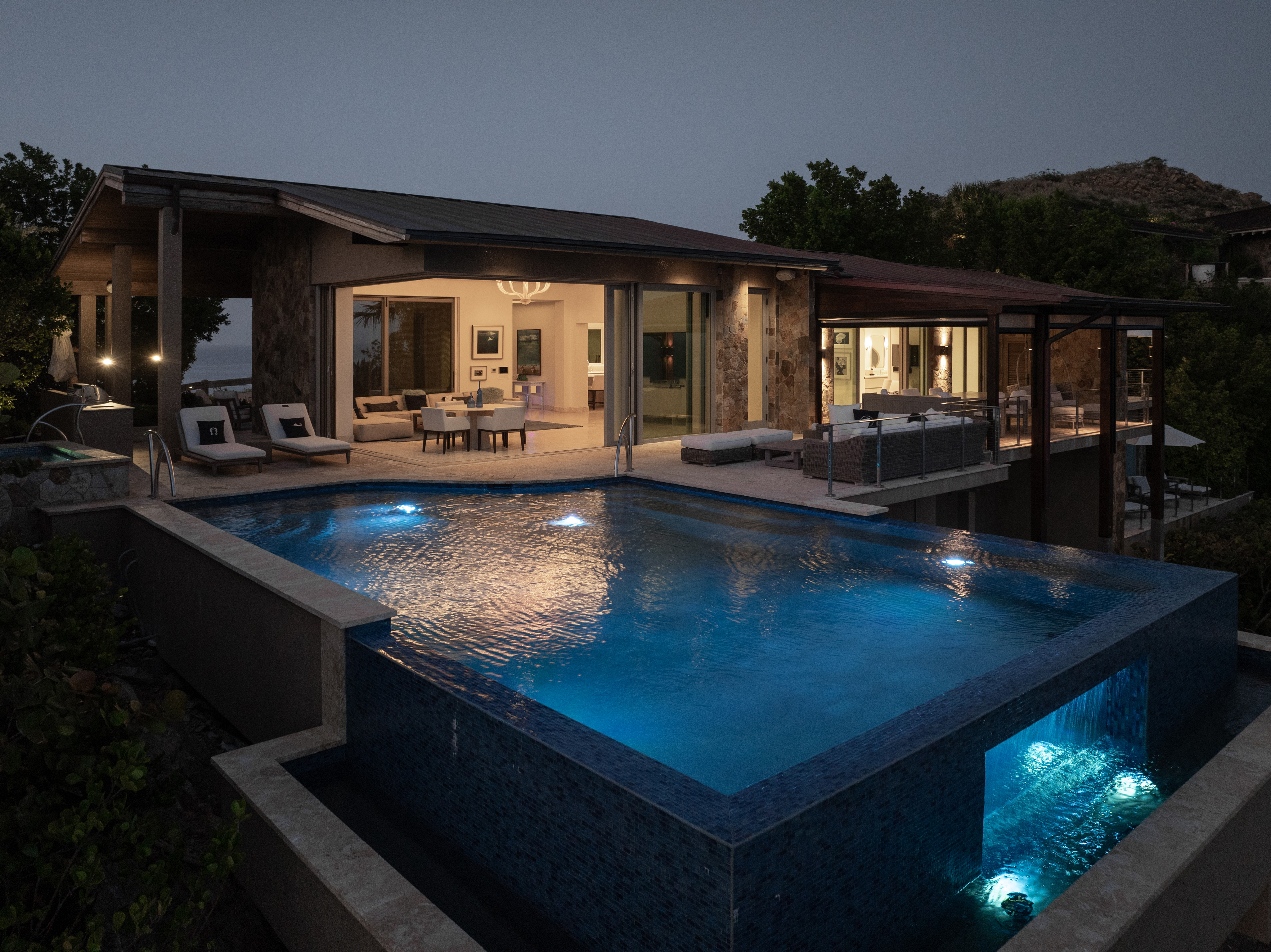 The Wild Side Villa at dusk features expansive glass walls and an infinity pool with underwater lights. The outdoor living area is illuminated, revealing contemporary furnishings. Surrounding trees and a distant hill complete the serene setting, making it the perfect villa rental for tranquility seekers.