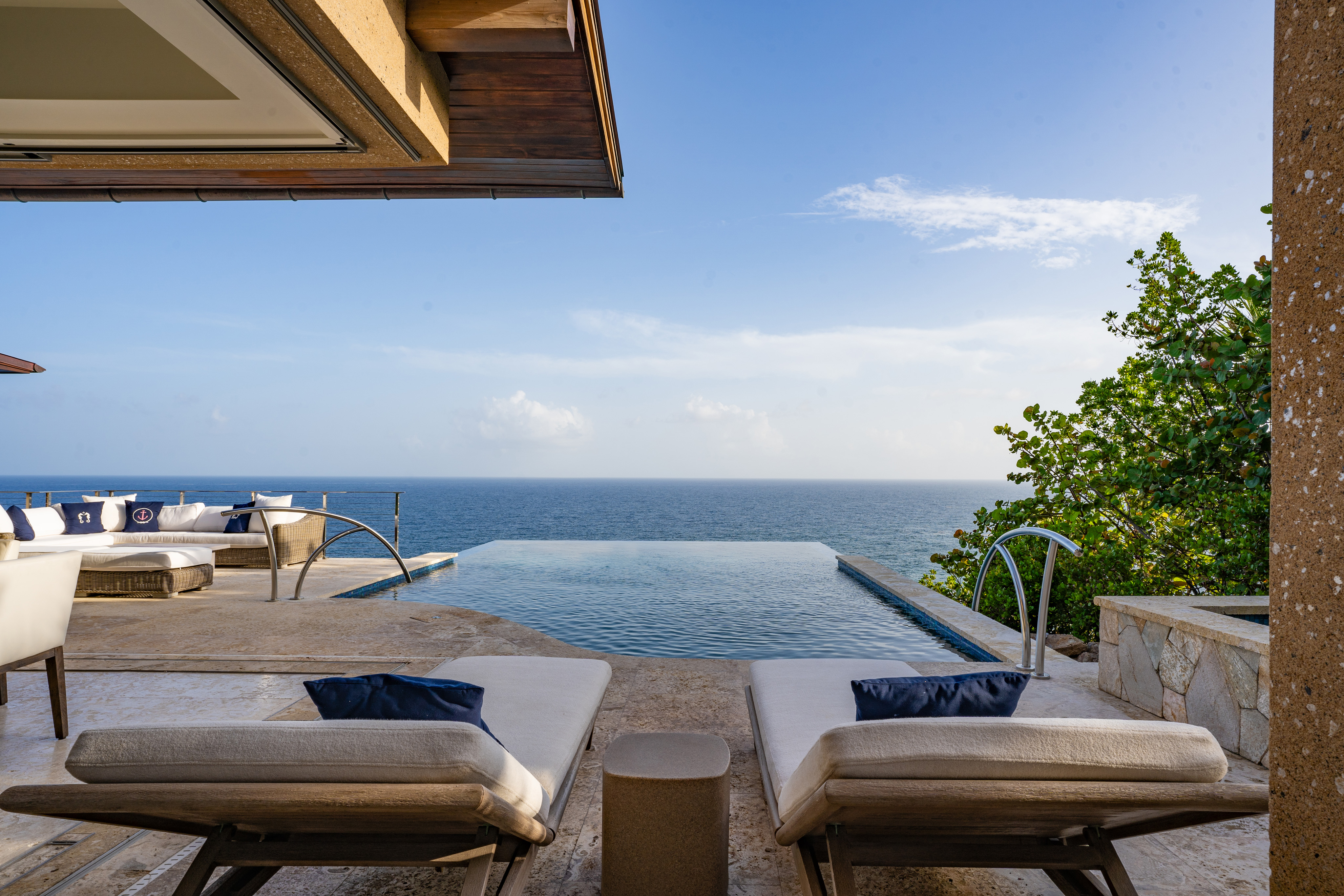 At Wild Side Villa, a luxurious infinity pool overlooks a serene ocean view. Two cushioned lounge chairs with blue pillows face the pool, positioned on a stone deck. To the left, a shaded seating area features a white cushioned couch and table. Lush greenery frames the scene.