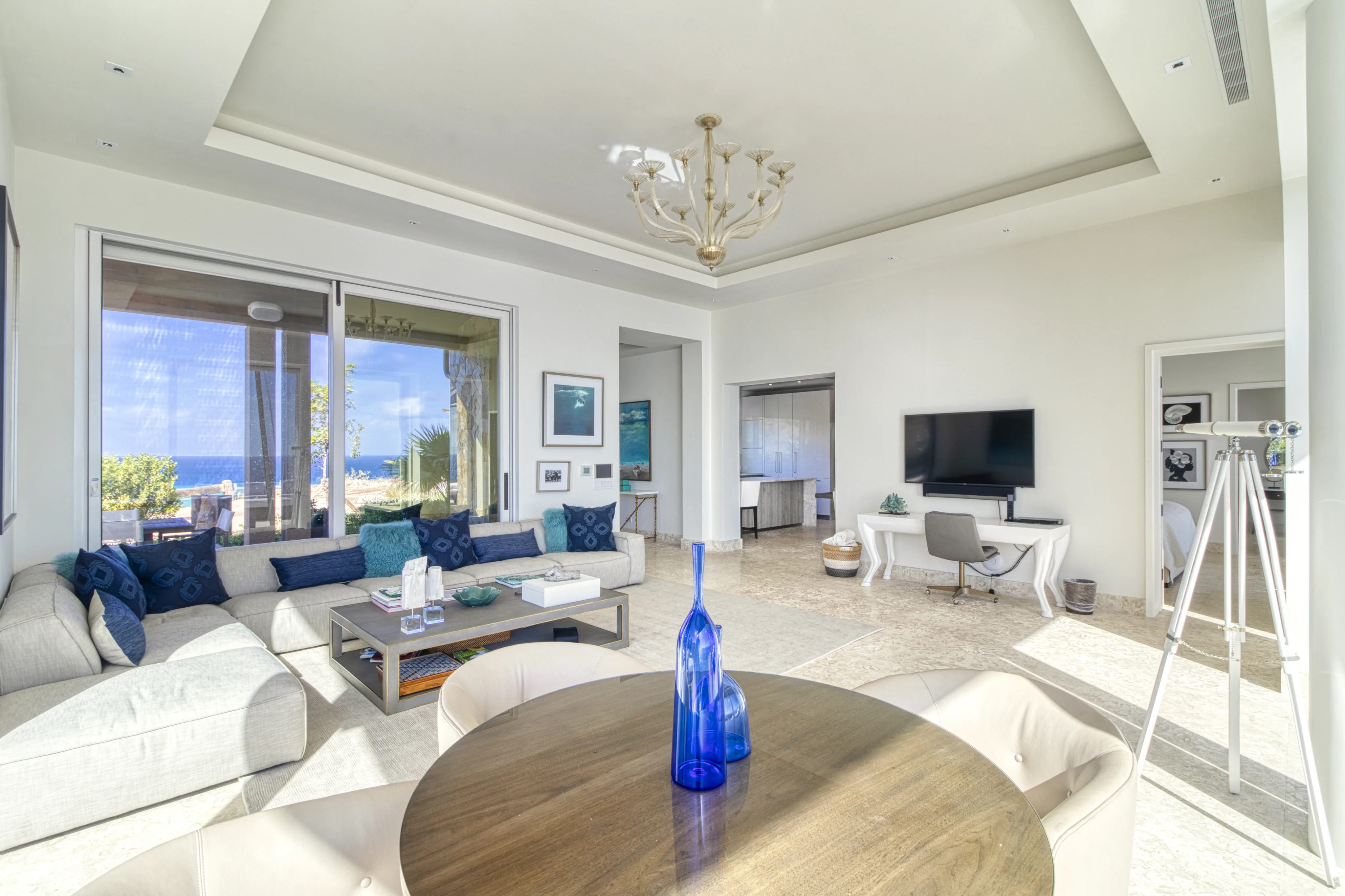 The bright, modern living room at Wild Side Villa features a large beige sectional sofa with blue cushions, a round wooden table with a blue vase, a wall-mounted TV, and a desk with a chair. Large windows offer an ocean view, and a chandelier hangs from the ceiling.