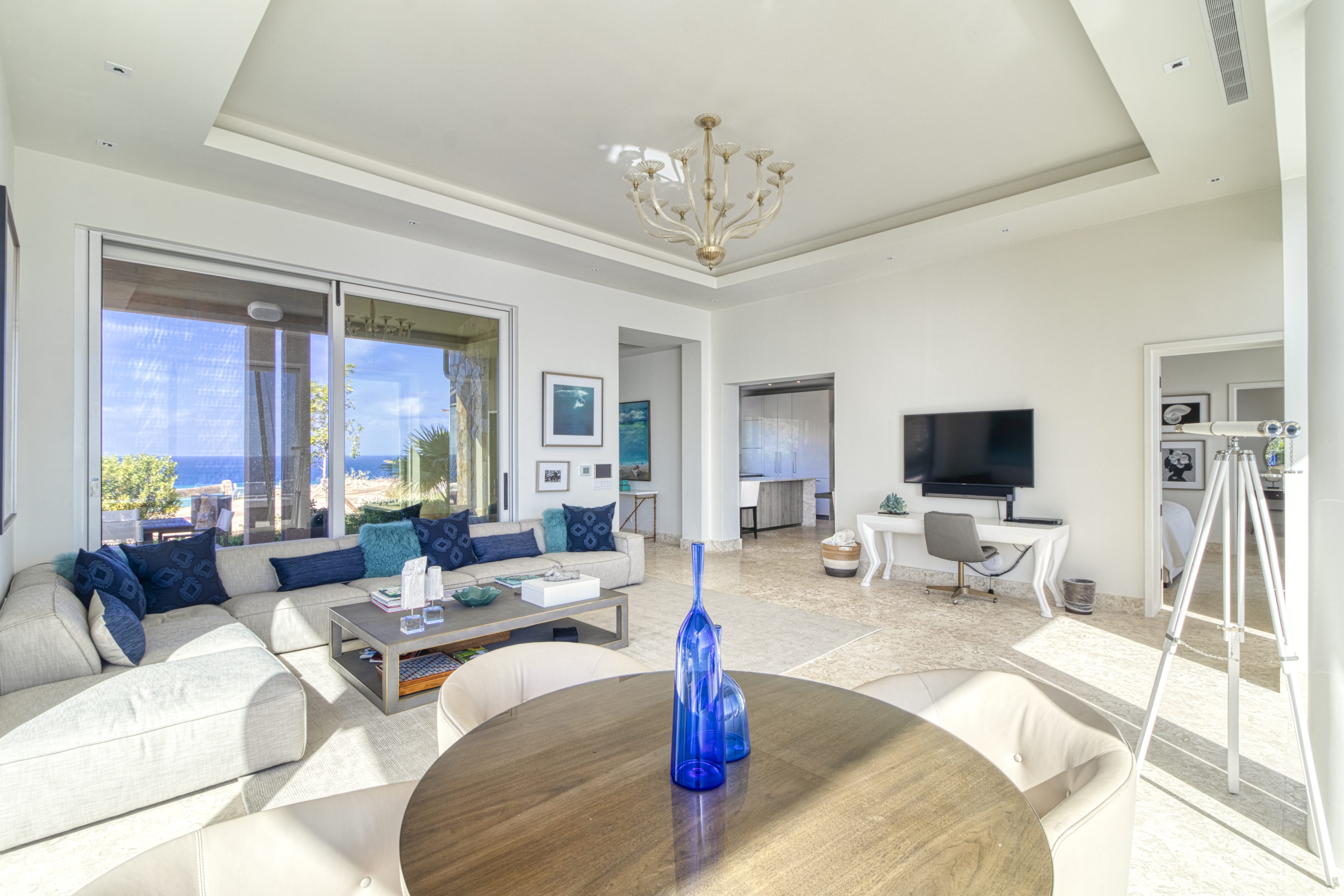 The bright, modern living room at Wild Side Villa features a large beige sectional sofa with blue cushions, a round wooden table with a blue vase, a wall-mounted TV, and a desk with a chair. Large windows offer an ocean view, and a chandelier hangs from the ceiling.
