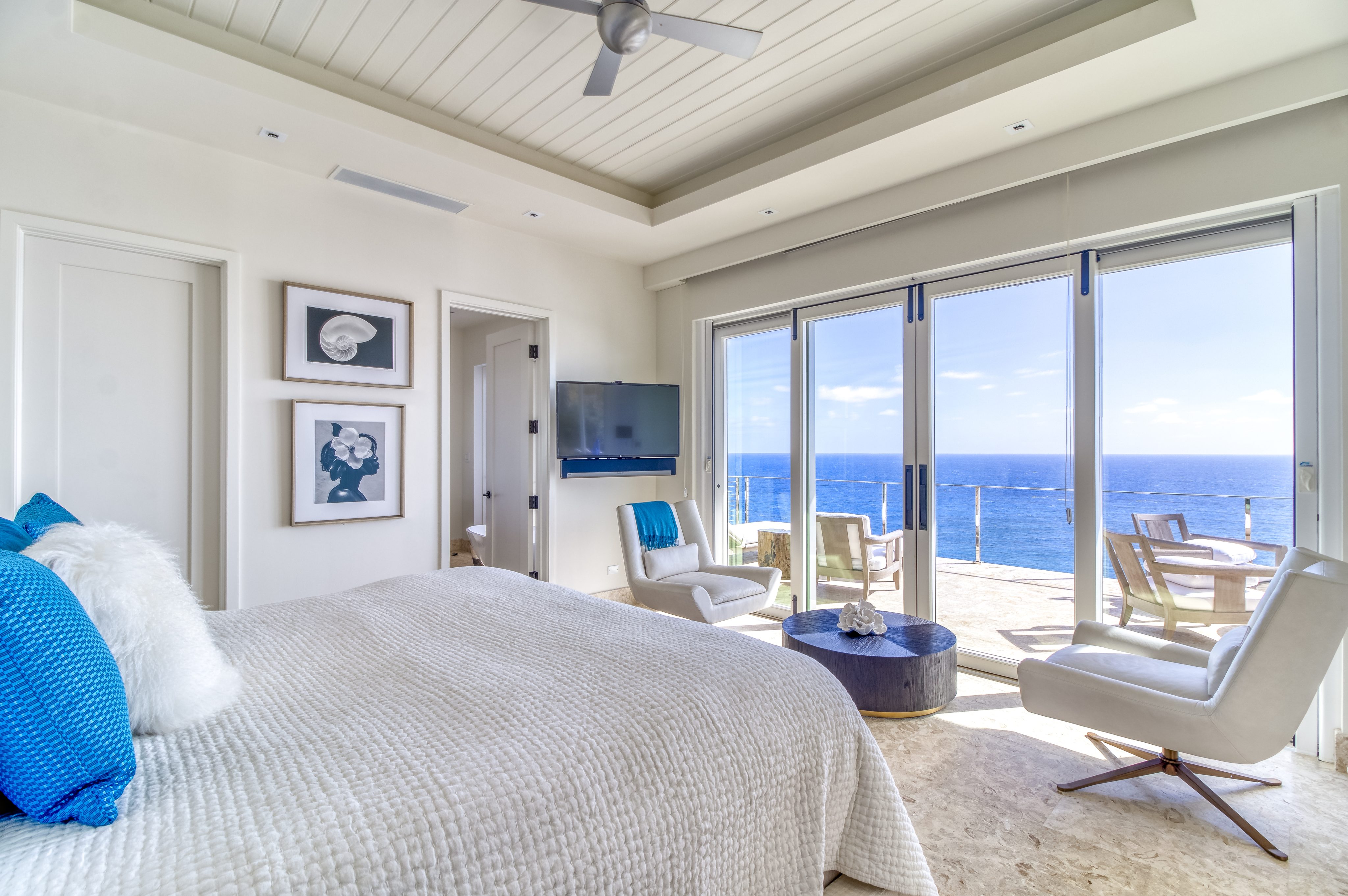 A bright and modern bedroom at Wild Side Villa featuring a large bed with white and blue bedding, a wall-mounted TV, and framed art. Floor-to-ceiling windows and sliding glass doors lead to a balcony with a seating area, offering a stunning view of the ocean.