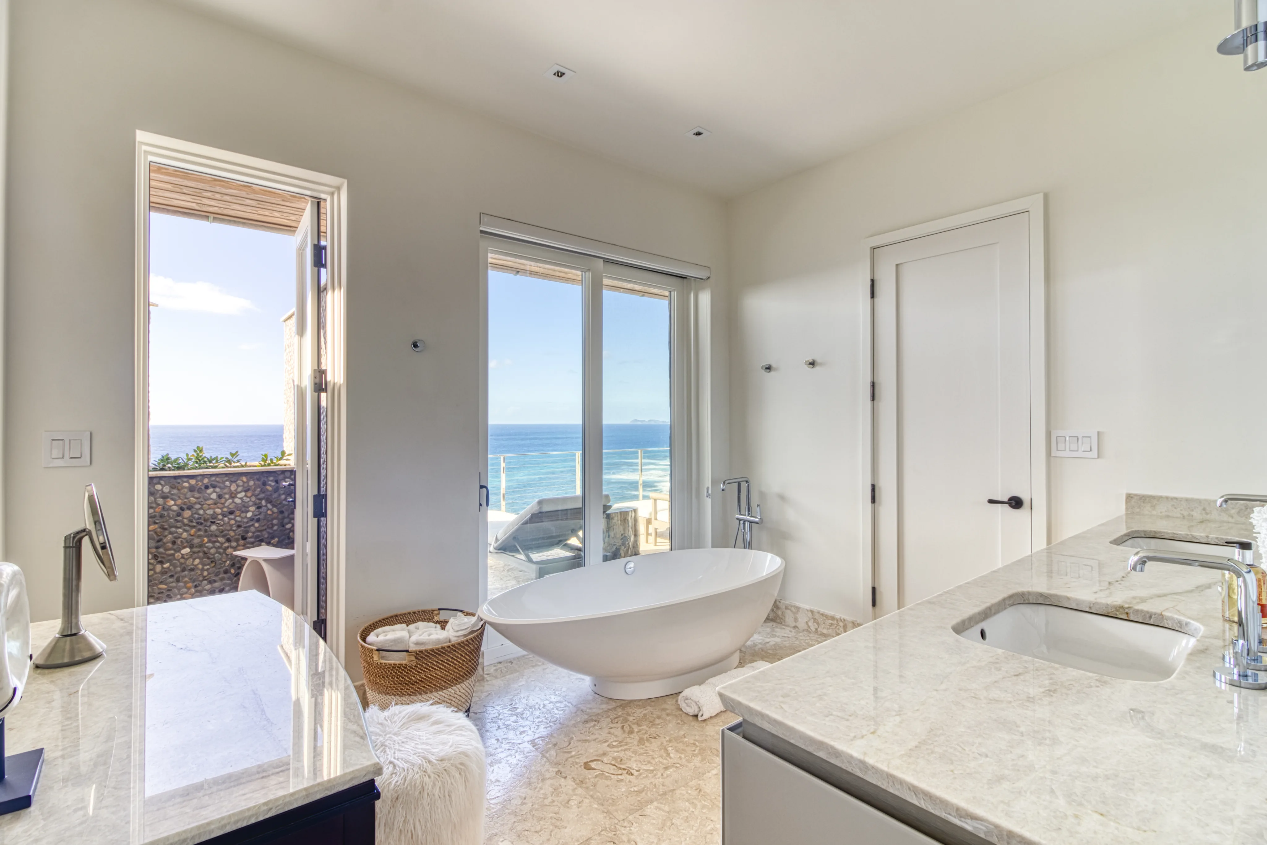 A modern bathroom at Wild Side Villa features a freestanding tub positioned in front of large windows offering an ocean view. The space includes marble countertops, a vessel sink, a fluffy white rug, and a wicker basket with towels. A door on the left leads to an outdoor area.