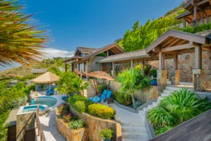 The image shows Poseidon's Perch Villa, a luxurious tropical retreat with a stone exterior nestled within lush greenery. There is a swimming pool with lounge chairs and thatched umbrellas, surrounded by beautifully landscaped gardens and a mountainous backdrop under a clear blue sky.
