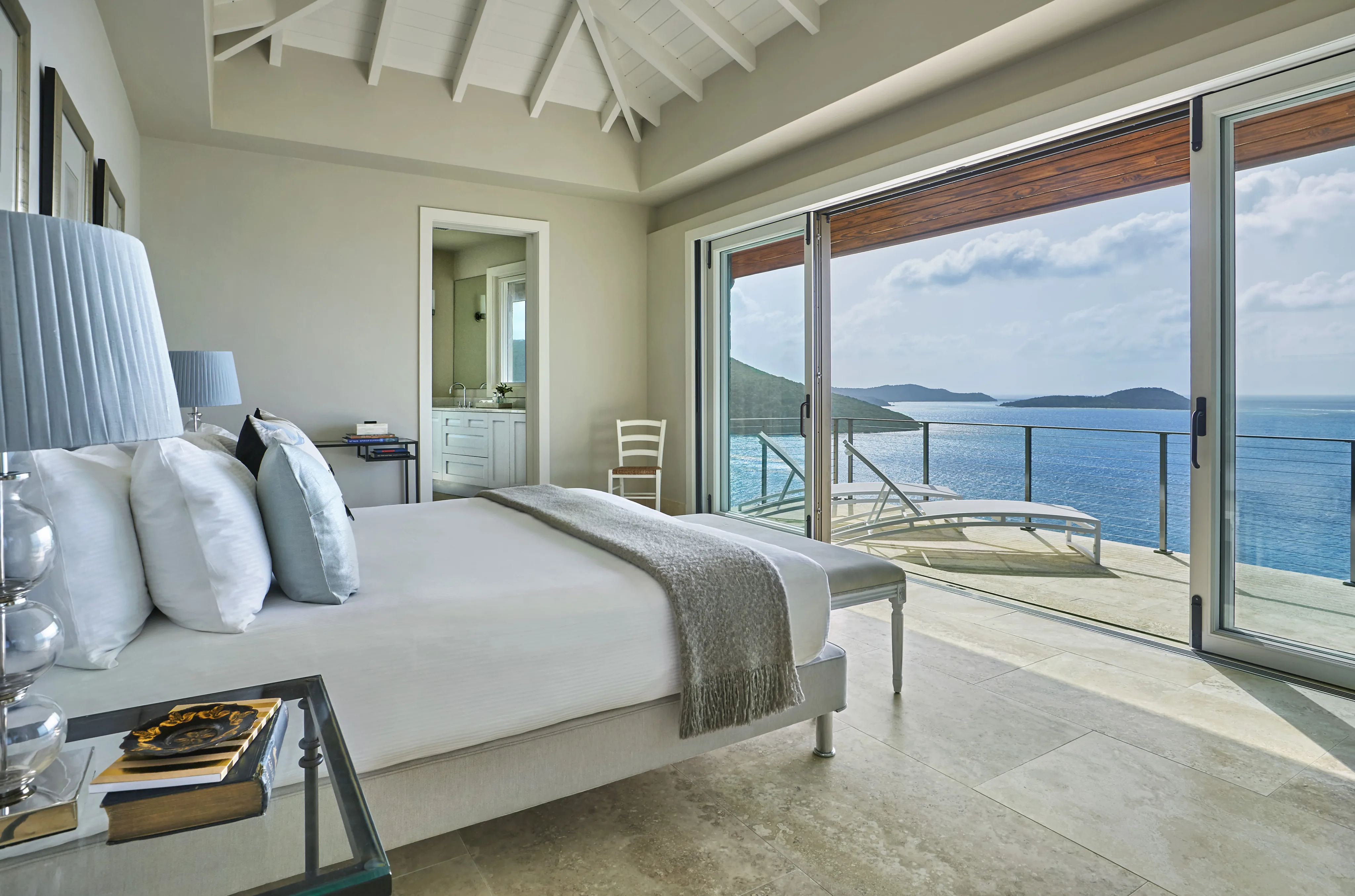 A bright, modern bedroom at Reef House features a large bed with white linens and a gray throw blanket. The room boasts a glass nightstand, a table lamp, and a white chair. Sliding glass doors open to a spacious balcony offering stunning ocean views, with the bathroom visible in the background.