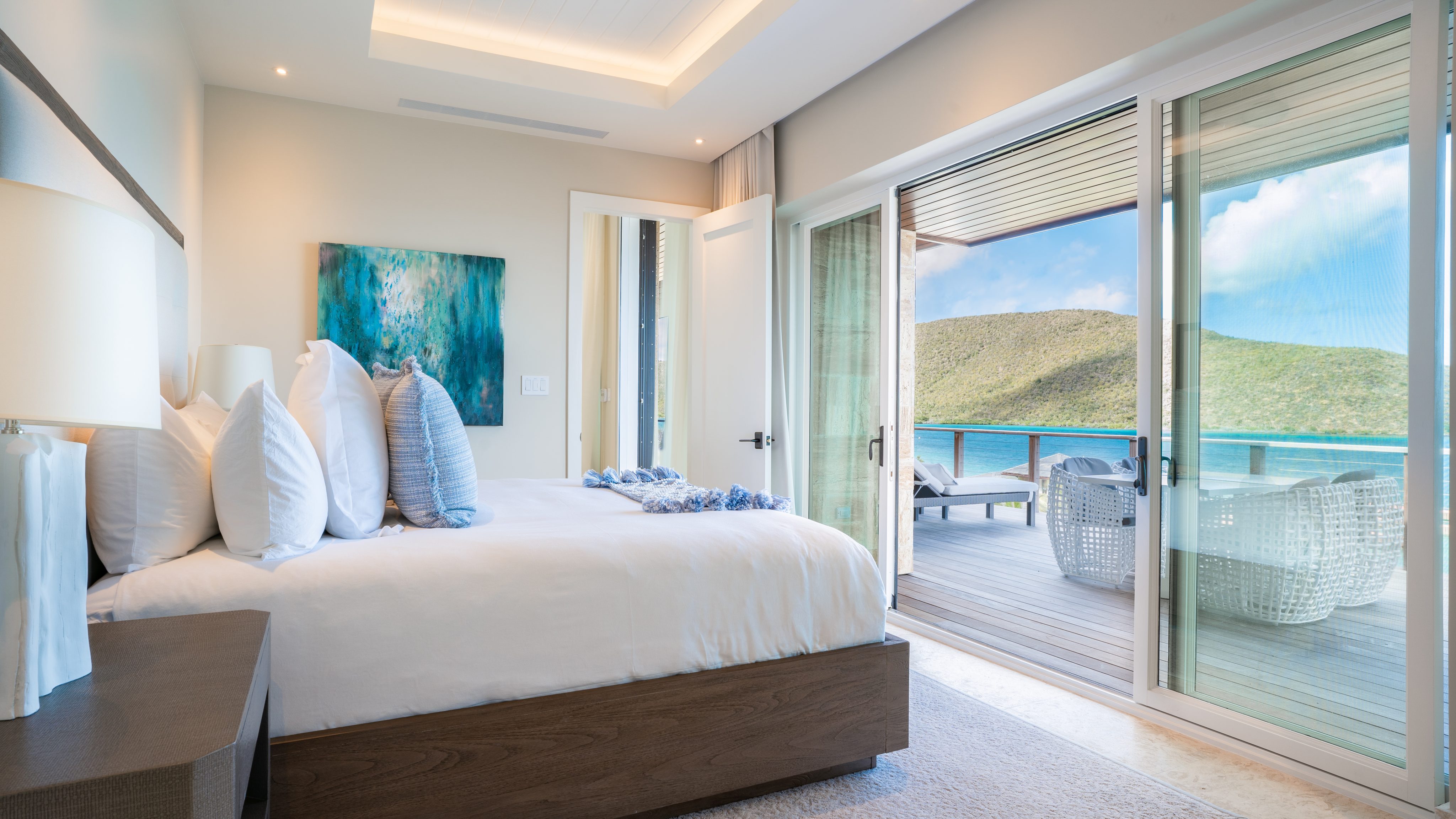 A bright, airy bedroom in Villa Sabella features a large bed adorned with white linens and blue accent pillows. A modern painting hangs on the wall beside a door leading to a spacious balcony. The balcony offers stunning views of a lush hillside and the turquoise sea under a clear sky.