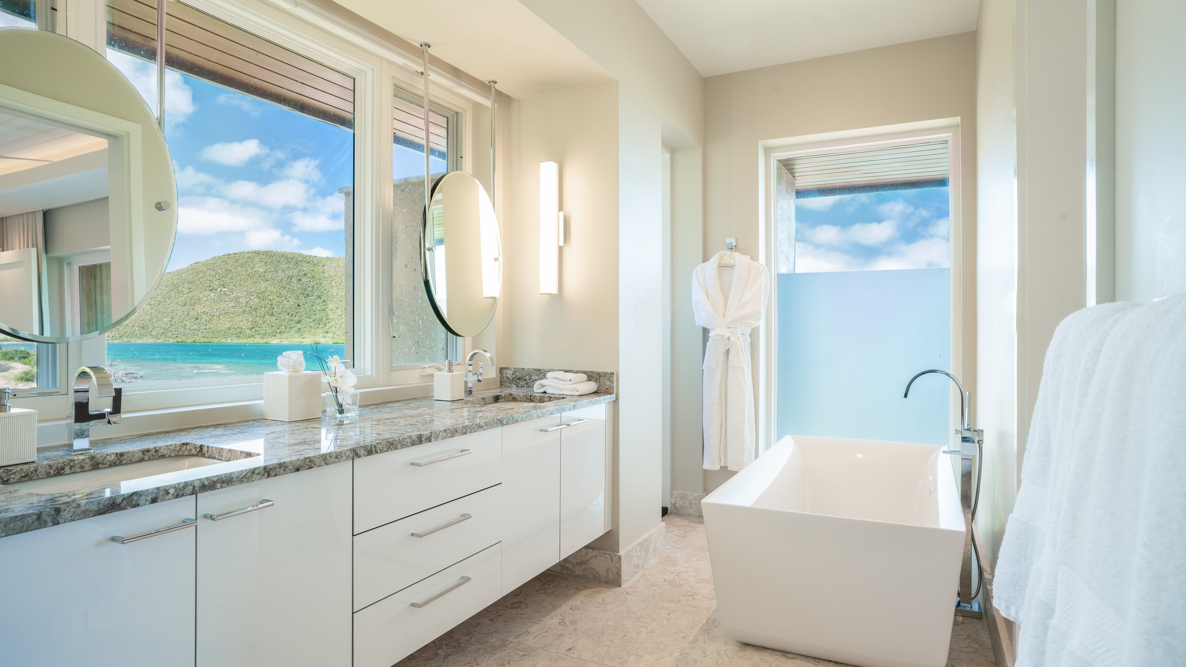A modern, luxurious bathroom in Villa Sabella with a large window overlooking a scenic view of green hills and a blue sky. The room features a double vanity with round mirrors, marble countertops, a freestanding bathtub, a towel rack with a white robe, and sleek white cabinetry.