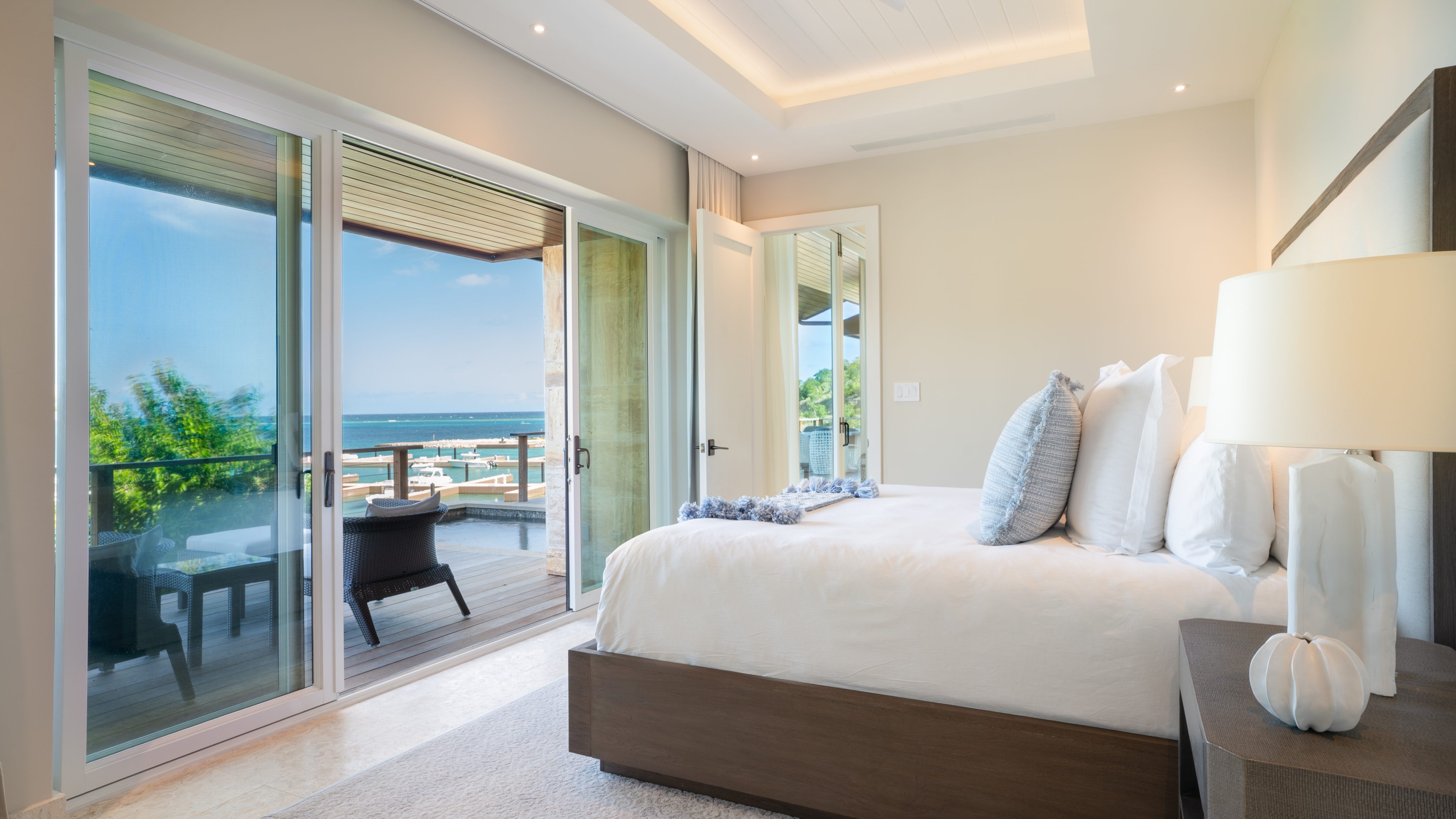 A serene bedroom in Sea Sabella Villa features a plush bed with white linens, flanked by a nightstand and lamp. Large sliding glass doors open to a balcony with seating, offering stunning ocean views framed by greenery under a clear blue sky. The room is softly lit and modernly furnished.