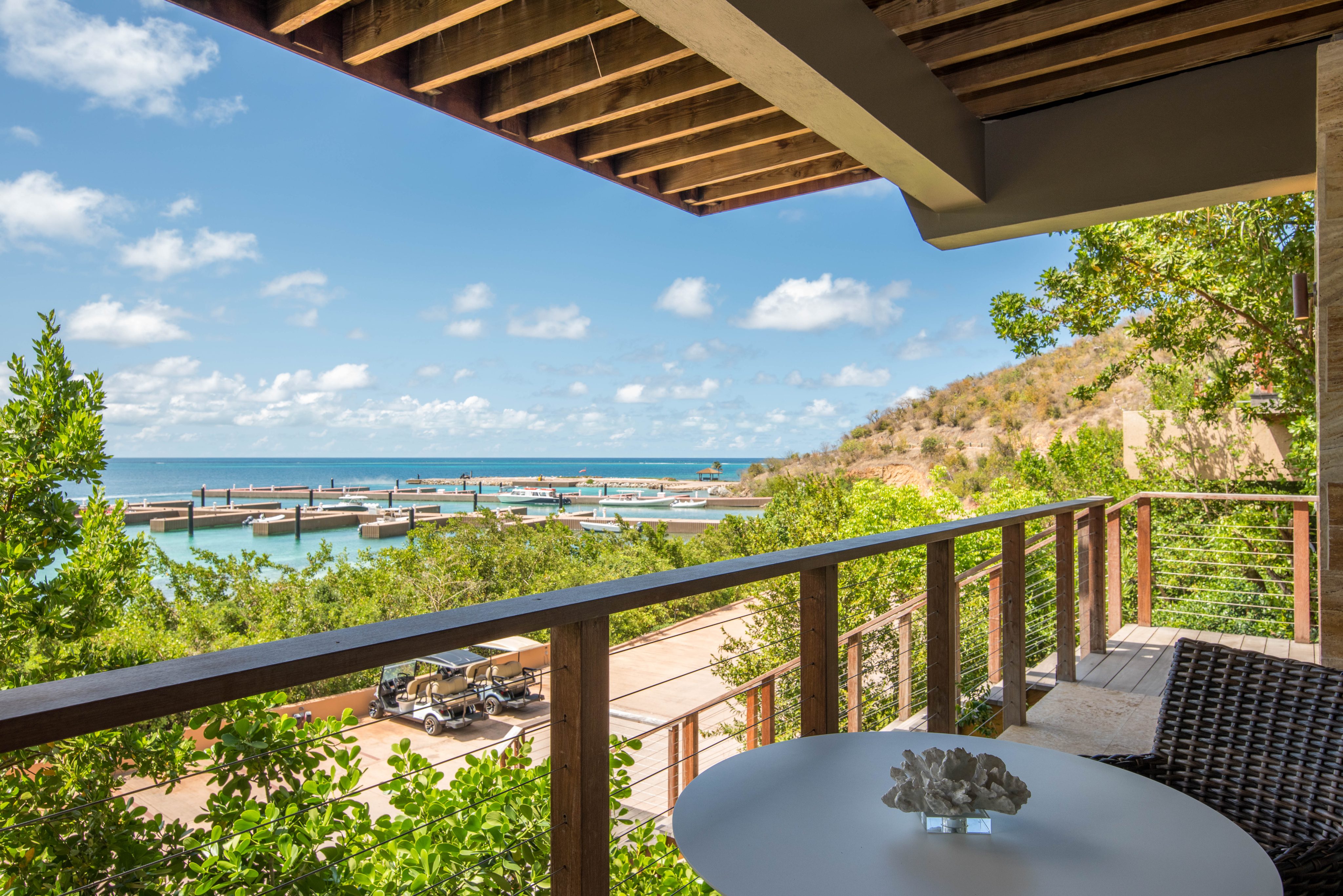 A serene view from Sea Sabella Villa's balcony overlooking a marina with clear turquoise waters and various boats docked. The foreground features a wooden table with a small decorative item, and lush greenery surrounds the scene under a partly cloudy sky.