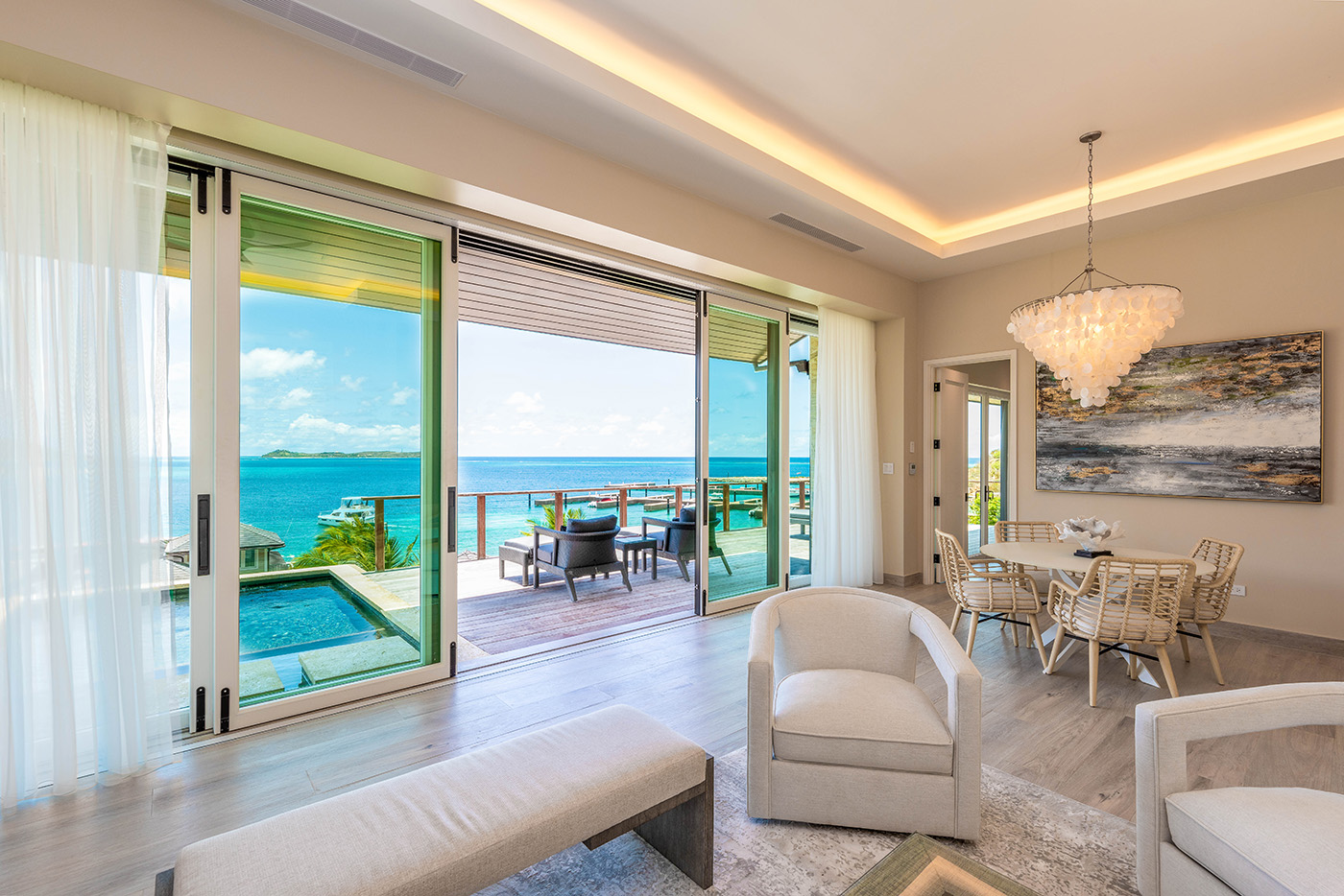 A modern living room in the exclusive Sur Villa features an open layout with a seating area, chandelier, and dining table. Large sliding glass doors lead to a patio with a pool and stunning ocean views. Light wood flooring and neutral tones create a bright and airy atmosphere.