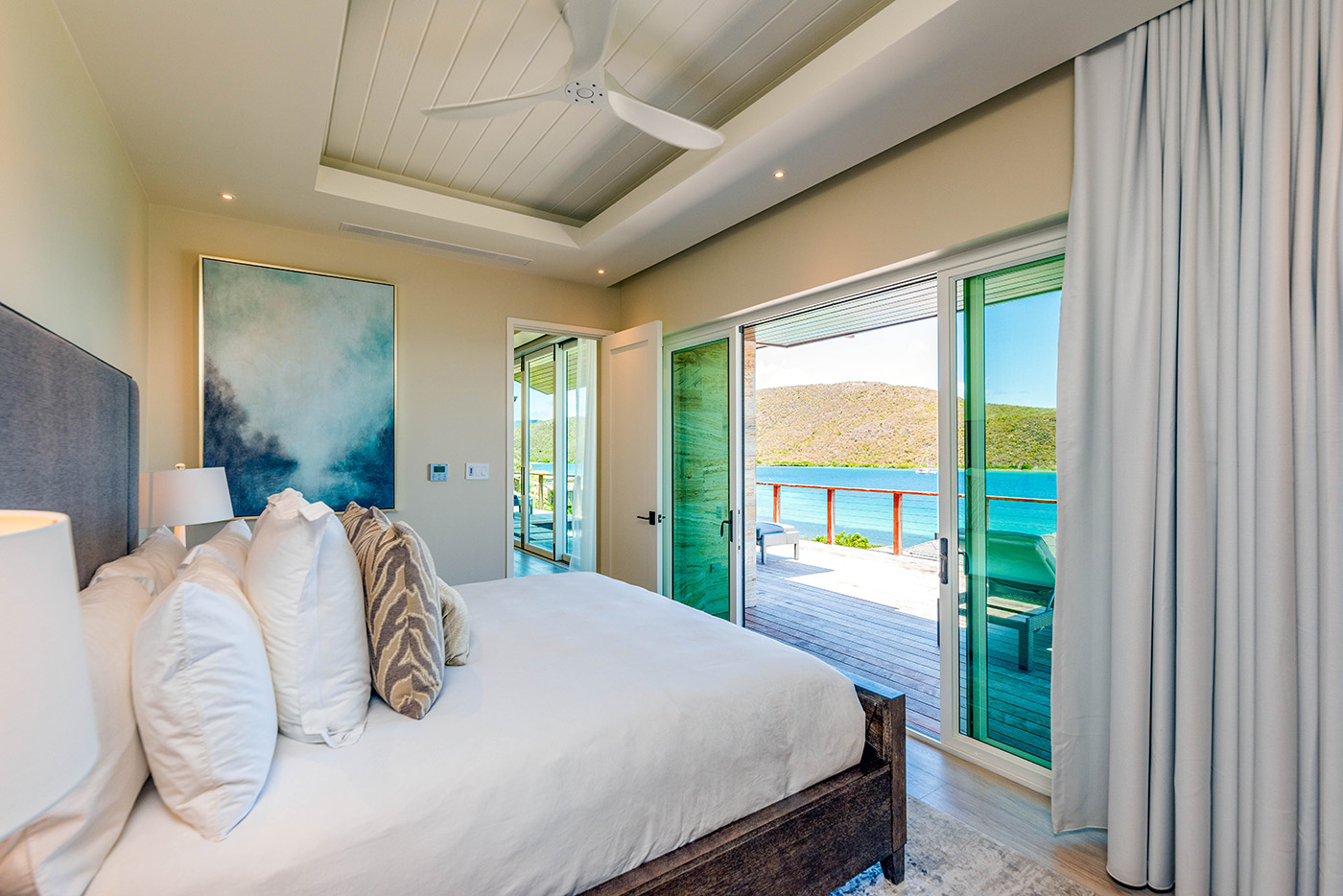 A modern Sur Villa bedroom features a large bed adorned with white and patterned pillows, a ceiling fan, and a beach-themed painting. Glass doors open to a balcony offering a scenic view of the ocean and hills. Light curtains frame the doors, and natural light fills the room.
