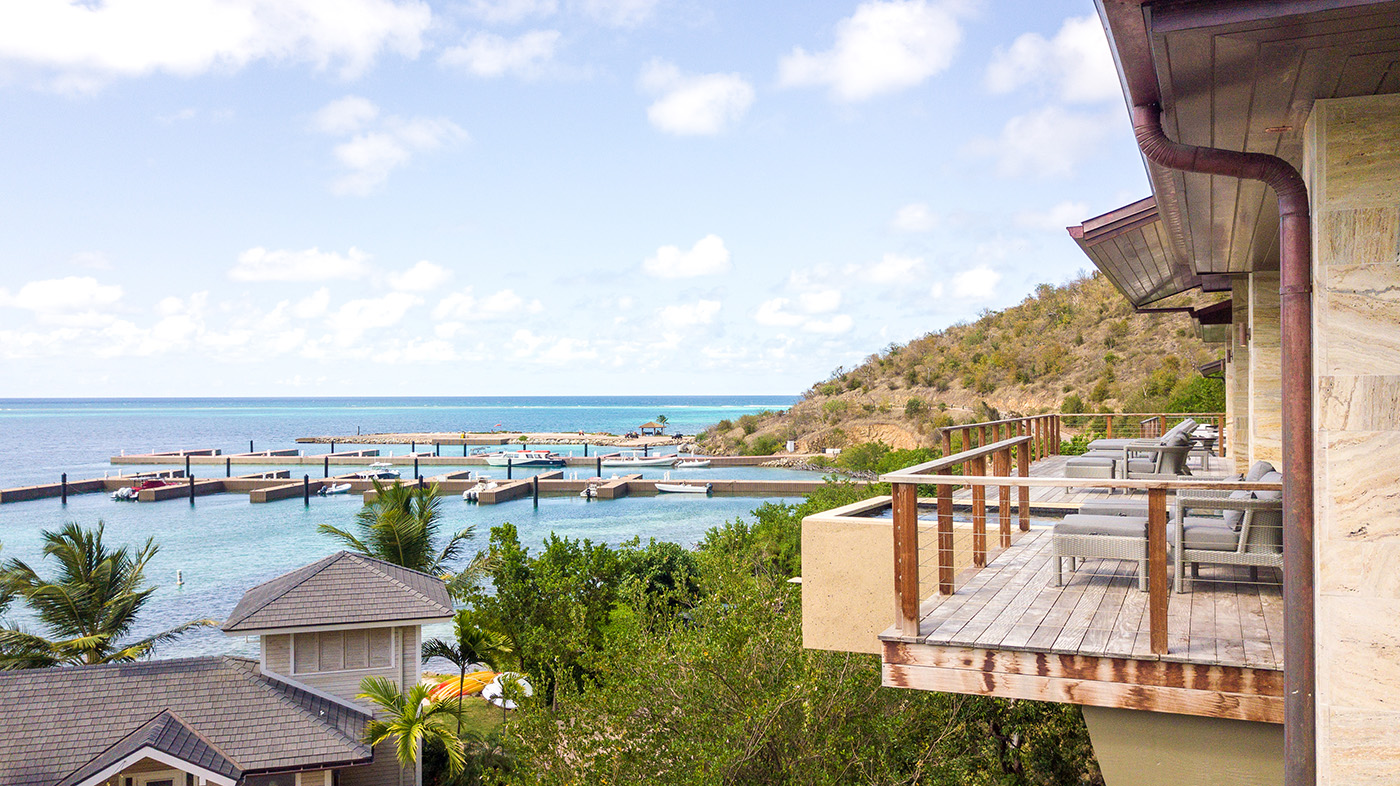 A scenic view of a coastal area featuring a wooden balcony with lounge chairs, offering a luxurious vacation experience overlooking the body of water with docks and boats. Lush greenery and a hillside are in the background, under a partly cloudy sky.