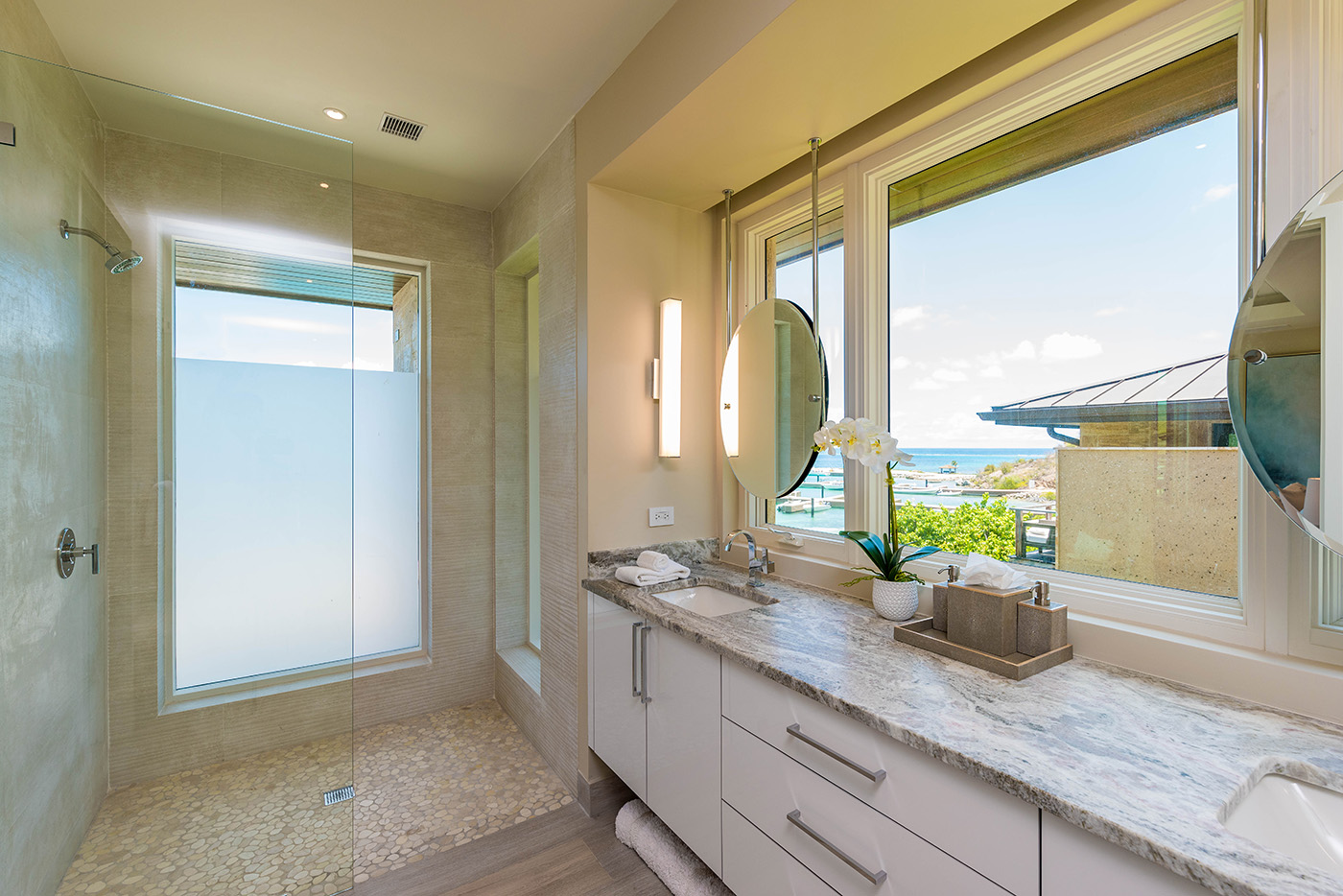 Modern Sur Villa bathroom with a large frosted glass shower, dual sinks, and a granite countertop. Two round mirrors adorn the space above the sinks. The room is well-lit with natural light from large windows showcasing a breathtaking view of the ocean.