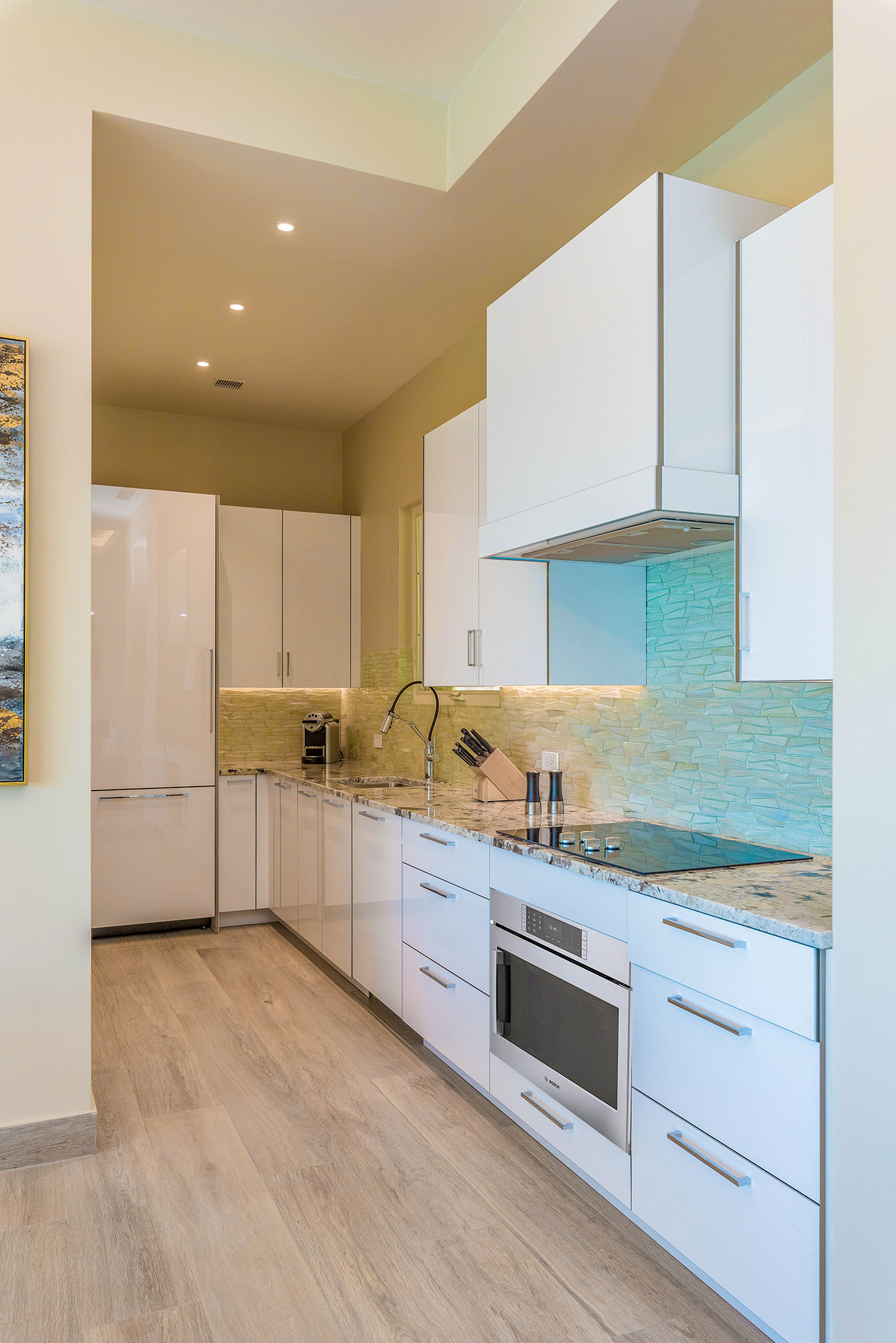 A modern kitchen in Sur Villa with white cabinets, light wood flooring, and a sleek design. It features stainless steel appliances, including an oven, stovetop, and range hood. The backsplash is a textured light green tile, and the countertops are granite.