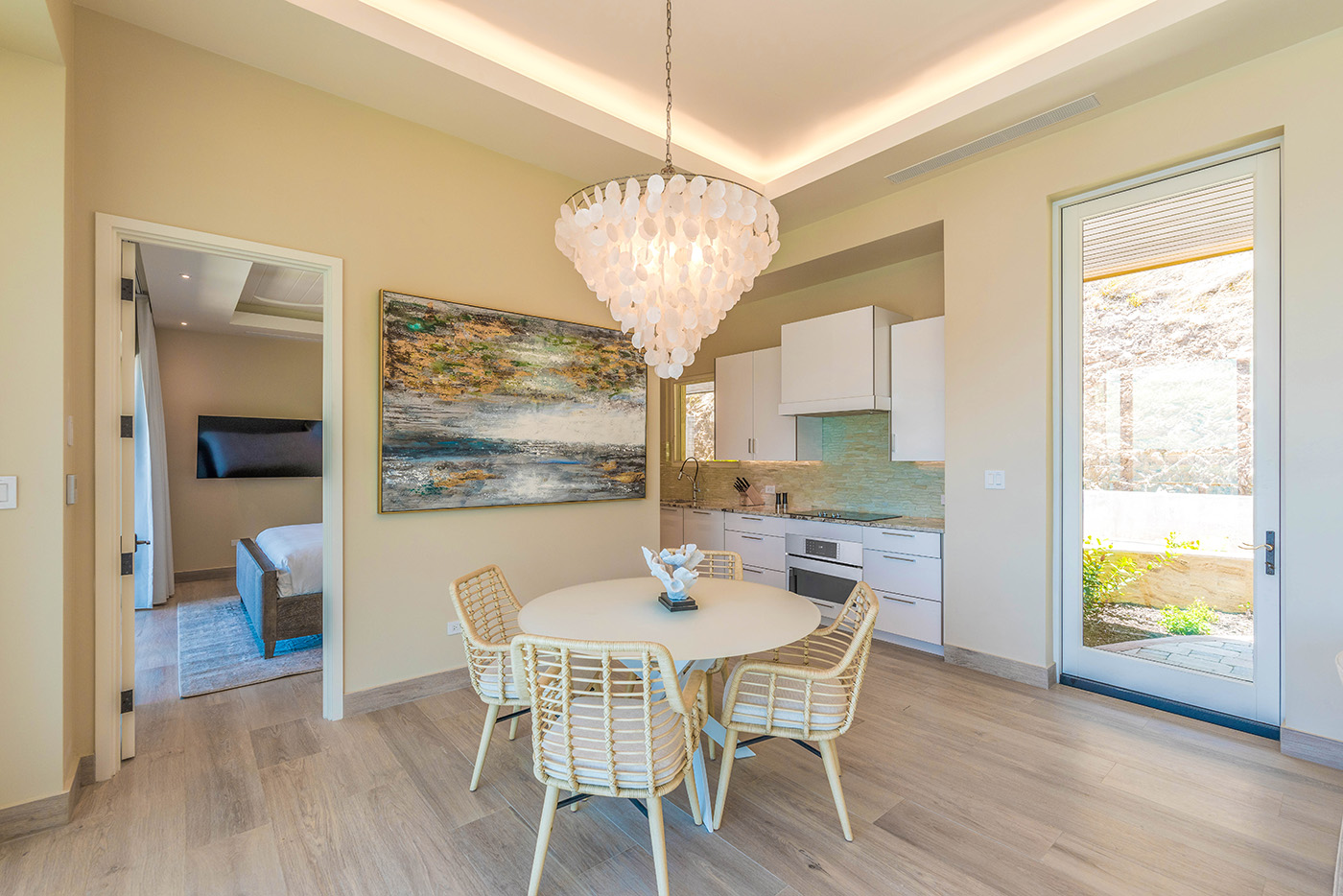Modern open-plan dining area in the villa featuring a round table with four wicker chairs under a large, decorative chandelier. A colorful abstract painting adorns the wall, with the kitchen and a doorway to a bedroom visible. Light wood flooring complements the neutral walls.