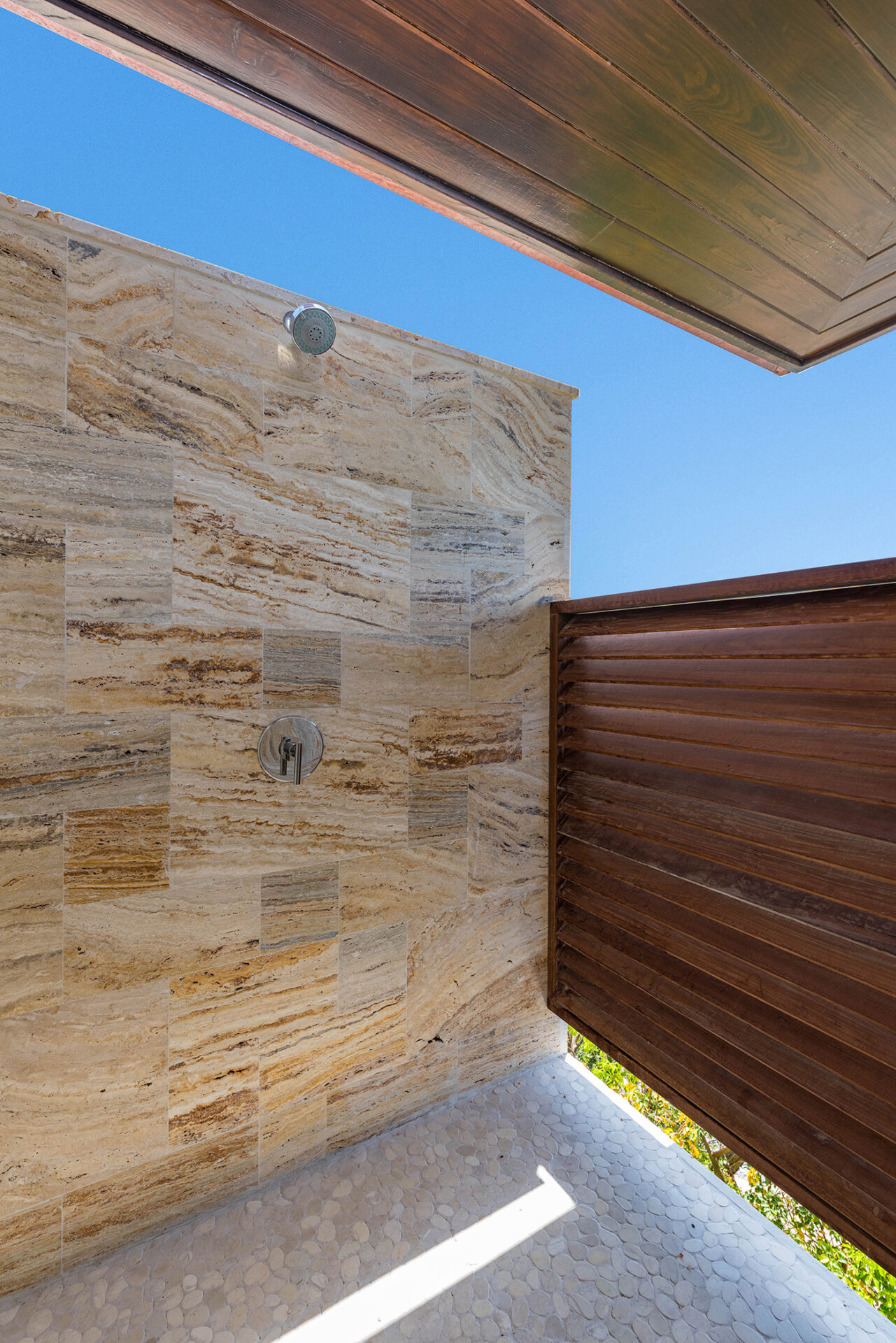 Outdoor shower with stone-tiled walls and a pebble-textured floor, surrounded by wooden slats offering partial privacy. The view from this Sur Villa gem opens up to a clear blue sky above.