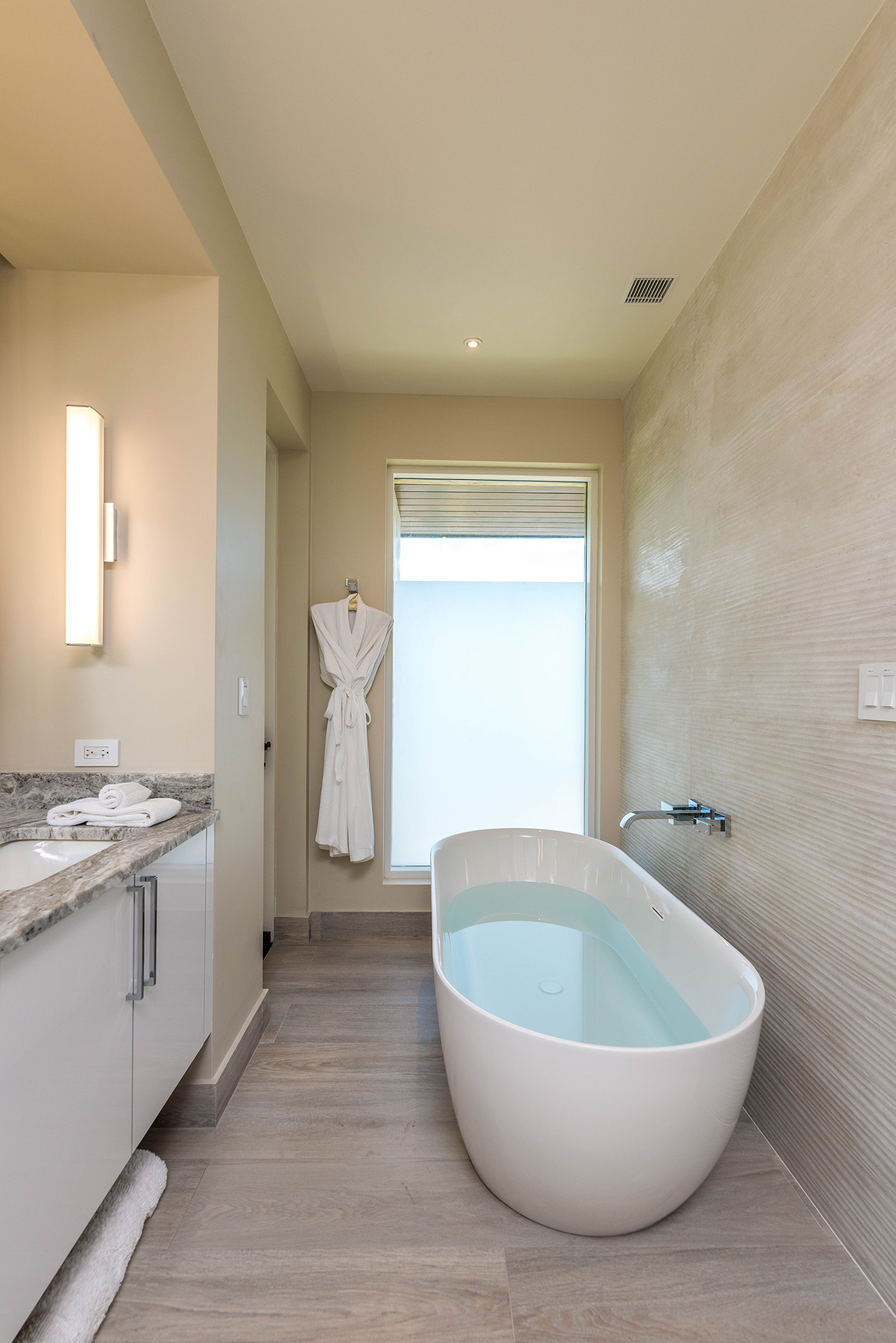A modern bathroom in Sur Villa features a freestanding white bathtub filled with water next to a beige tiled wall. A plush white robe hangs on a hook near a frosted window, while light-colored wood flooring and a white vanity with a granite countertop and wall-mounted light complete the serene space.