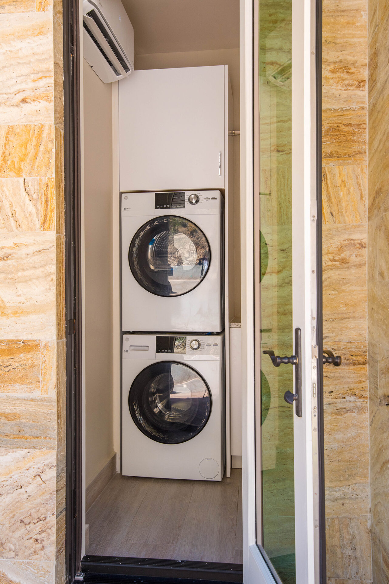 A modern laundry closet in a luxurious villa, featuring stacked white front-loading washer and dryer units. Enclosed by a glass door with a black handle, the walls boast textured beige and yellow tiles. Above the appliances, an air conditioner unit ensures comfort during your stay at this exquisite travel destination.