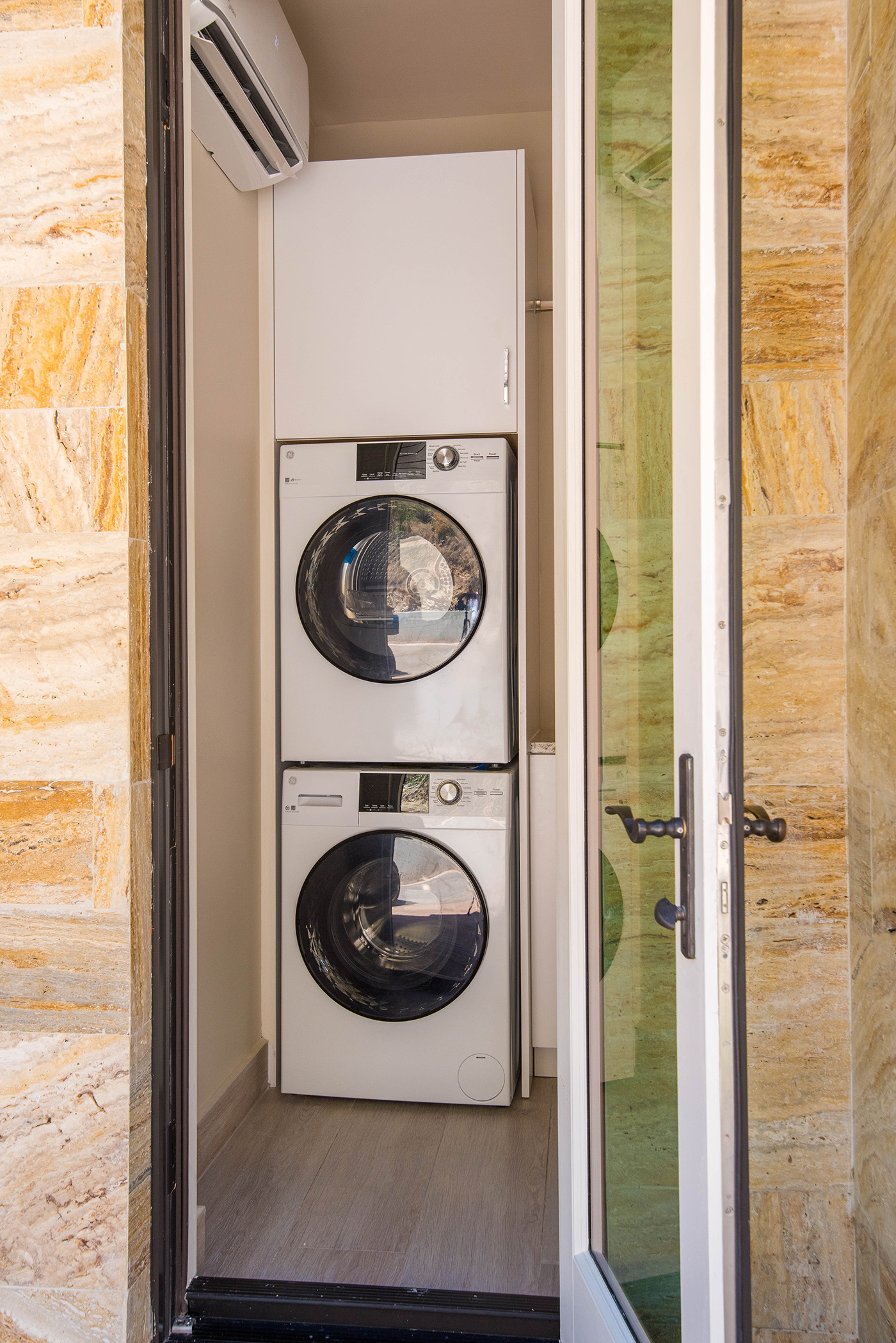 A modern laundry closet in a luxurious villa, featuring stacked white front-loading washer and dryer units. Enclosed by a glass door with a black handle, the walls boast textured beige and yellow tiles. Above the appliances, an air conditioner unit ensures comfort during your stay at this exquisite travel destination.