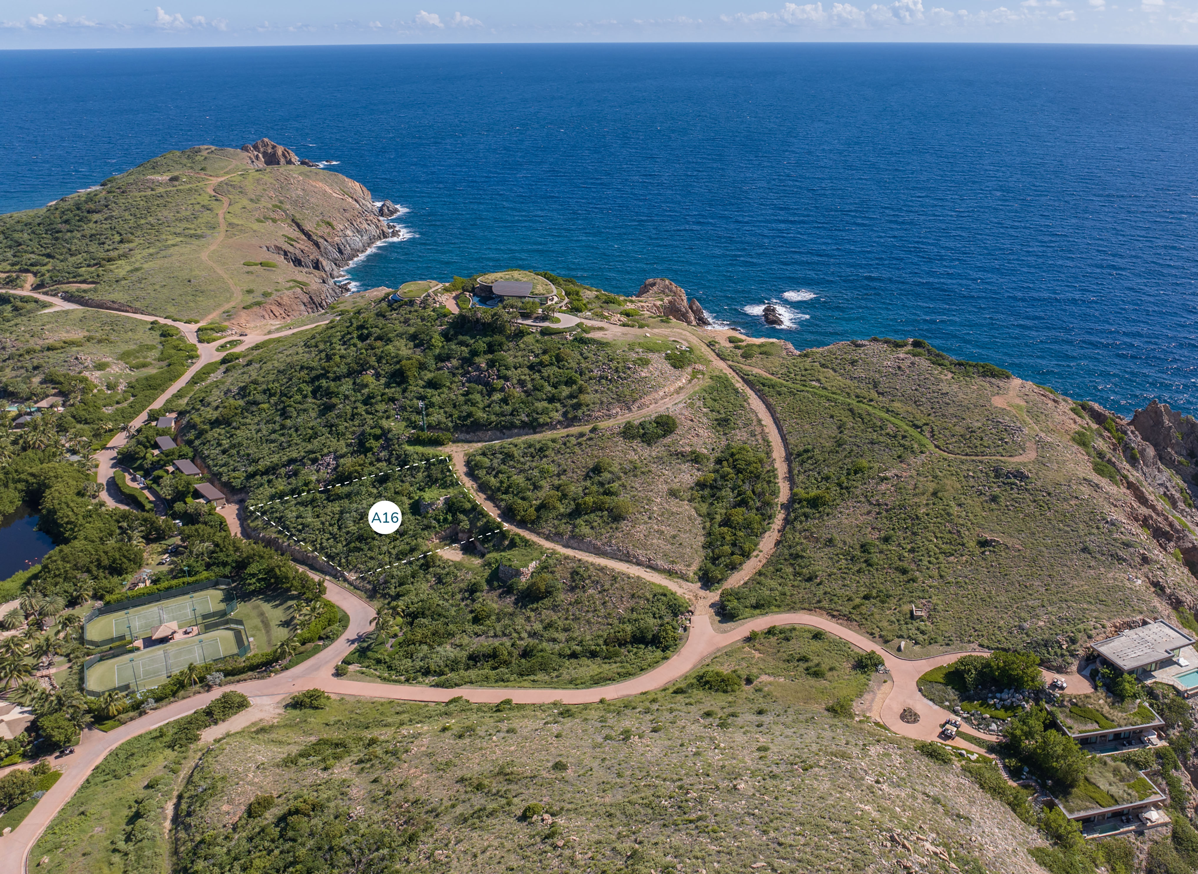 Aerial view of a coastal area with green hills and winding roads. There is a circular building near the Atlantic Ridge, tennis courts lower down, and a few scattered homesites. The ocean is visible in the background with a marker labeled "A16" on one of the buildings.