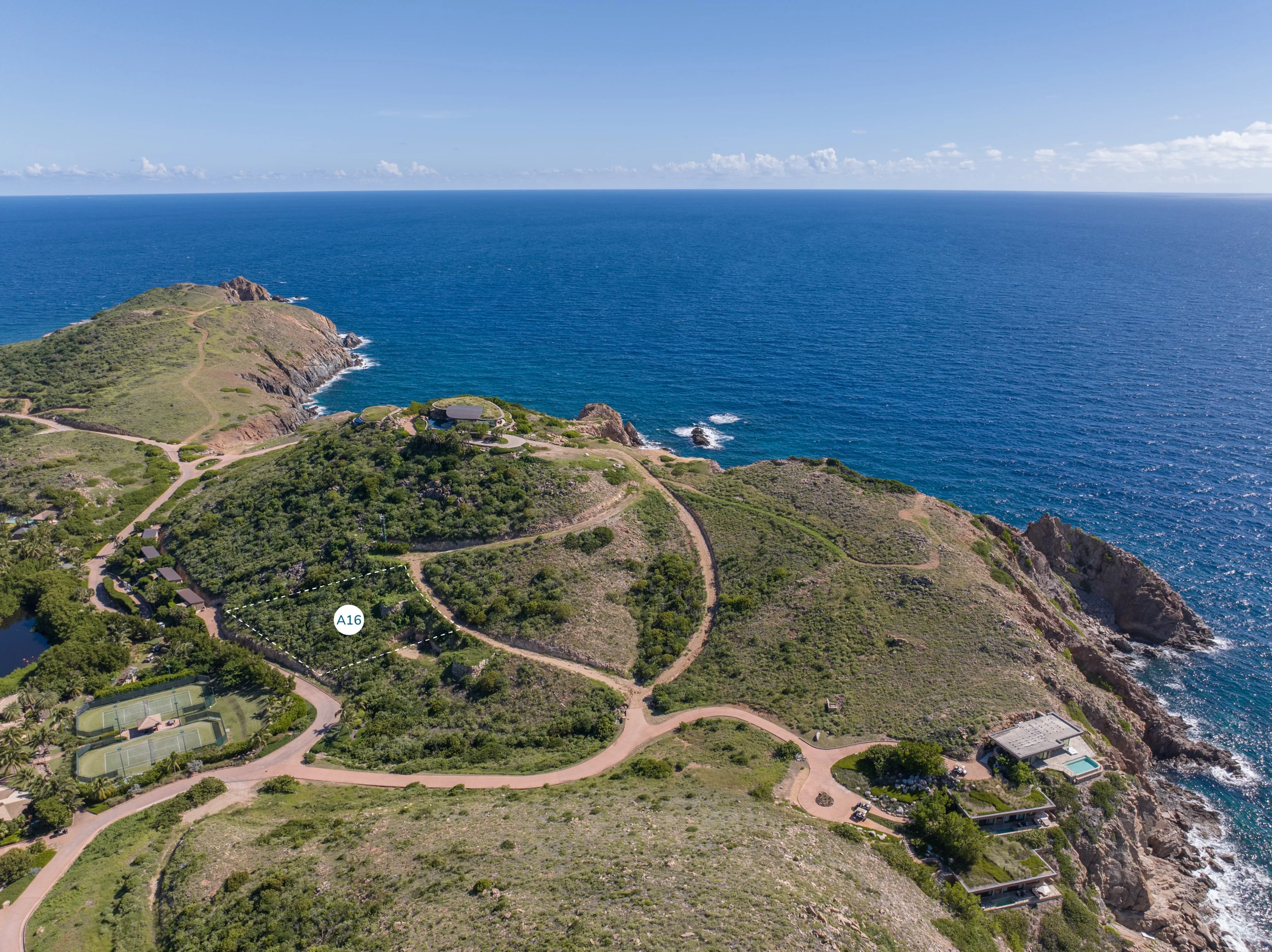 Aerial view of a rocky coastline along the Atlantic Ridge with lush green hills and winding dirt roads. The landscape features scattered homesites and pathways leading towards the sea. A calm blue ocean stretches into the horizon under a partly cloudy sky.
