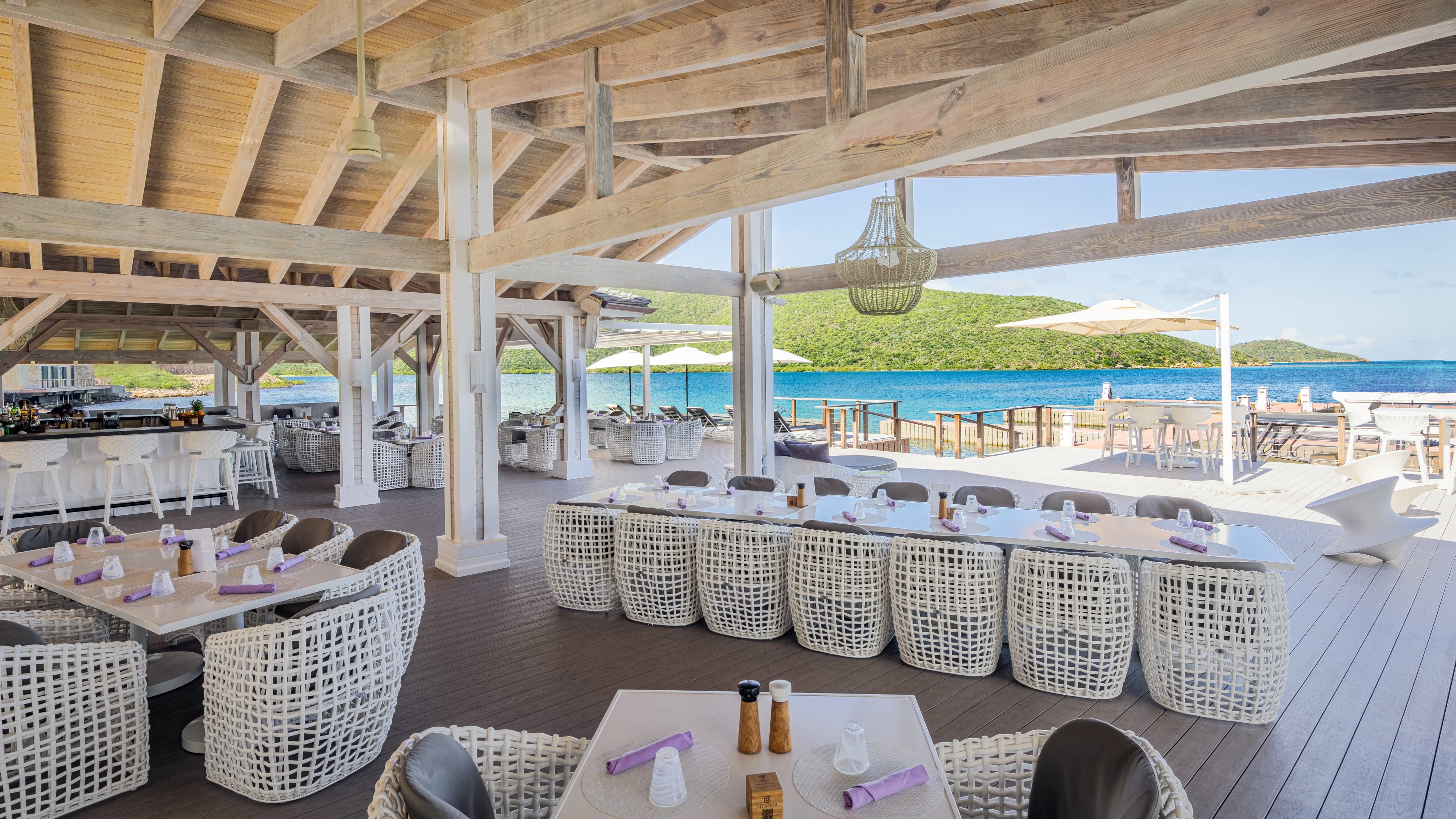 Open-air restaurant with wooden beams, wicker furniture, and neatly set tables overlooks a clear blue sea and green hills. White umbrellas provide shade on the sunny deck, enhancing the serene coastal ambiance. A well-stocked bar is visible in the background, making it one of the best things to do in the BVI.